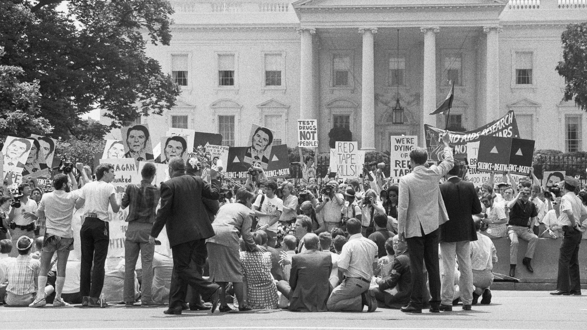 Black and white image of a protest in front of the White House with signs with Ronald Reagan's face and sayings "Silence equals death."