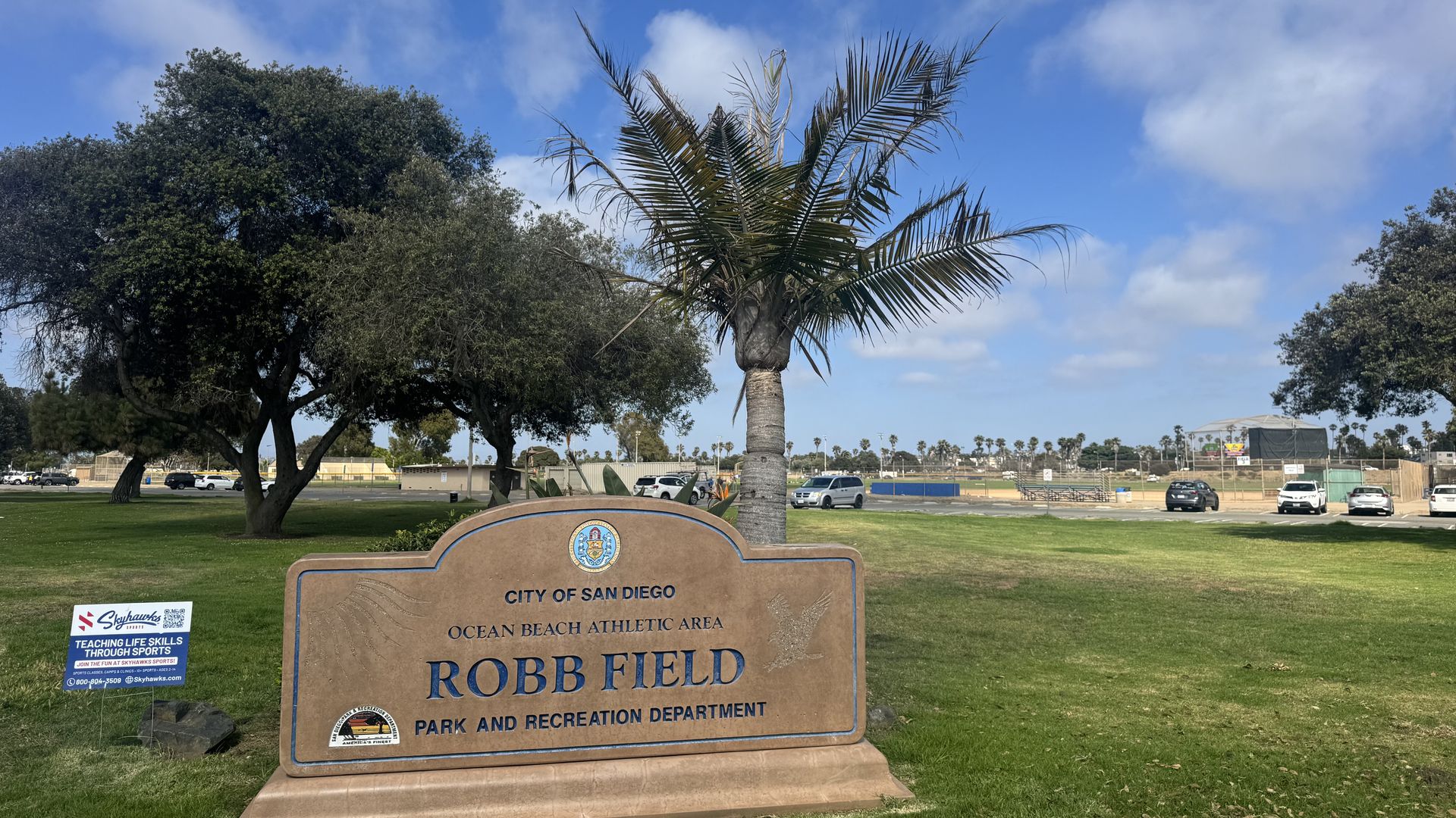 The entrance of Robb Field with a sign and palm tree, and cars parked in the background.