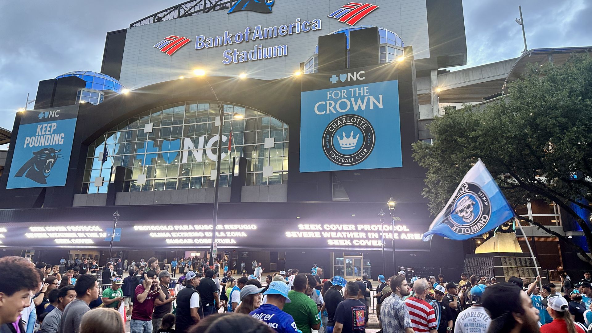 Exterior view of Bank of America Stadium with the Carolina Panthers logo at the top, flags on the roof, blue banners reading FOR THE CROWN, and a large crowd of fans outside in team colors.