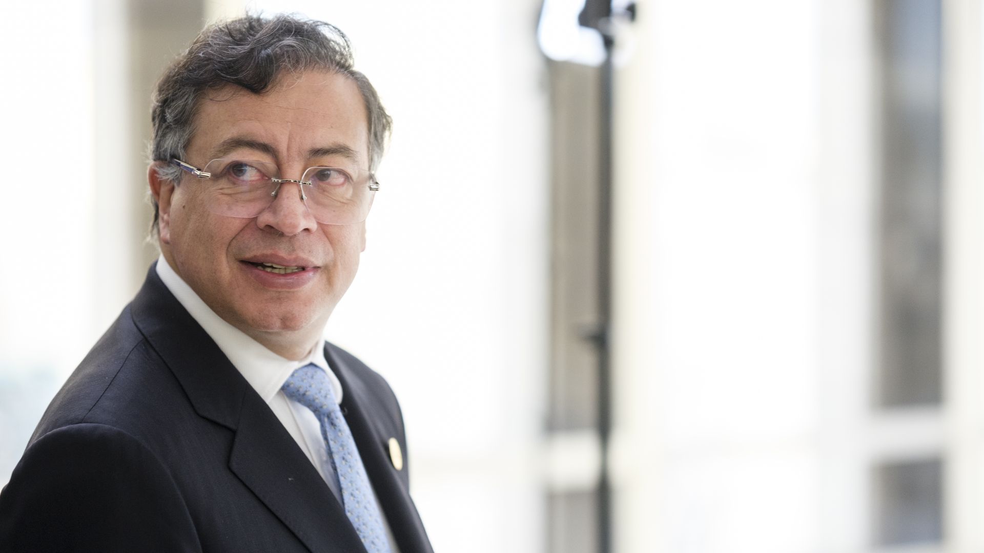 A man with a suit, white shirt and blue tie, wearing glasses, looks to his right.