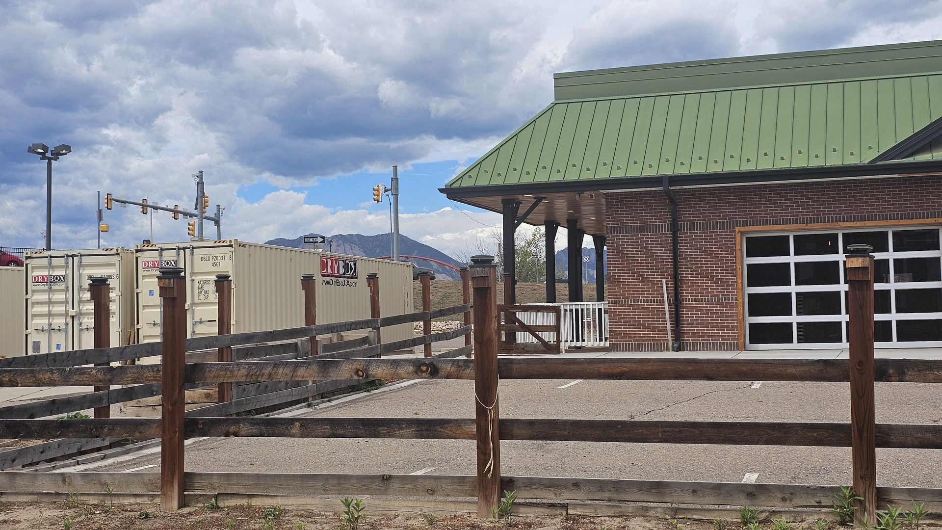 Brick building with a green sloped metal roof on the right, beige shipping containers labeled DRY BOX behind a wooden fence, and a cloudy sky with distant mountains.