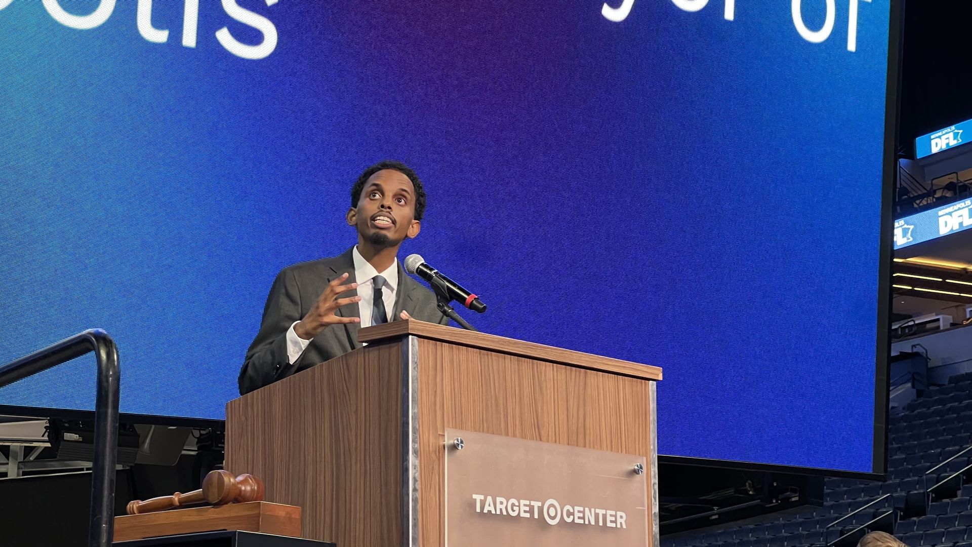Omar Fateh, wearing a dark suit, speaks passionately at a wooden podium labeled "TARGET CENTER" with a large blue screen behind him and a gavel nearby.