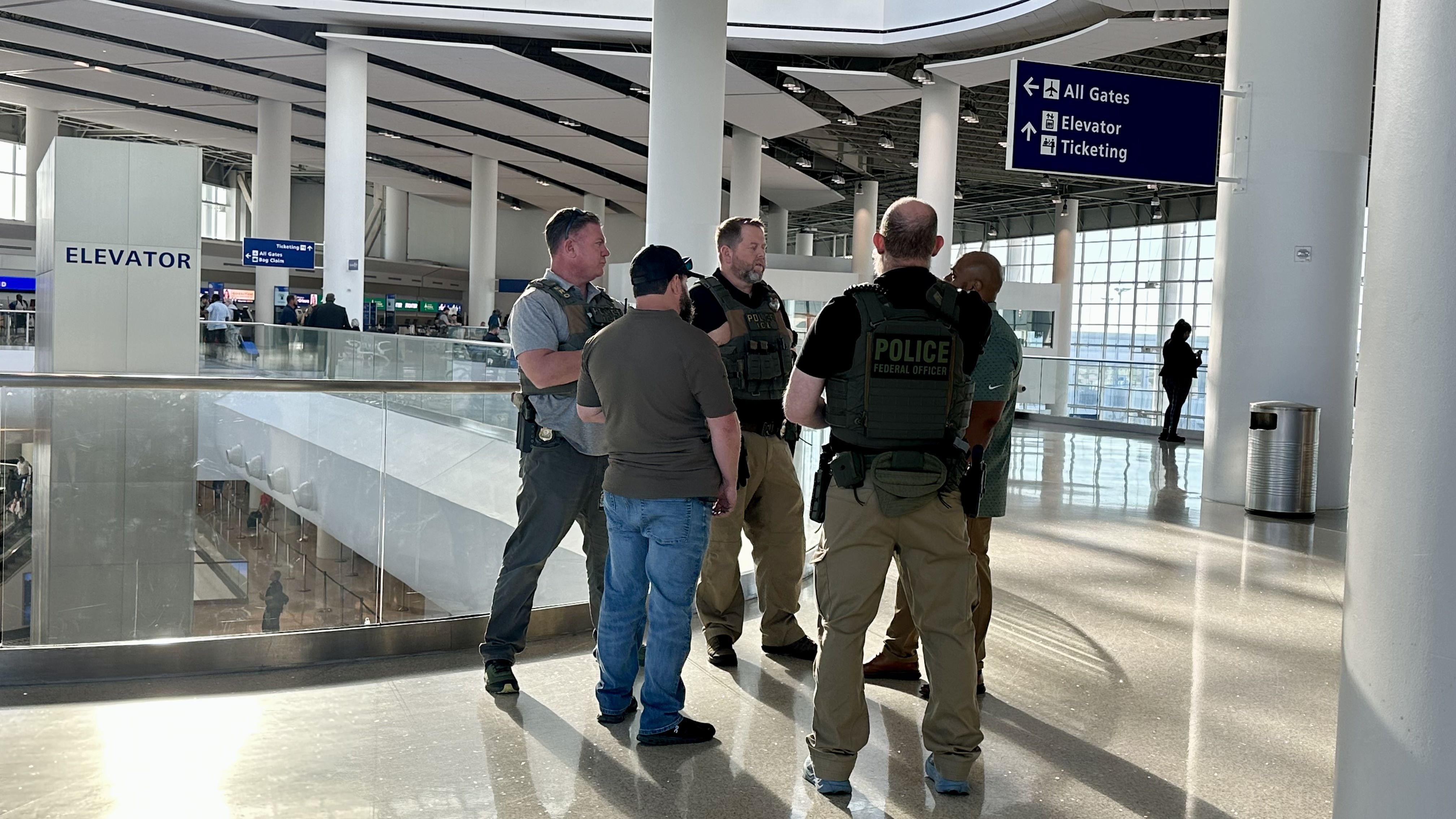 Five men in tactical vests chat in a bright airport terminal near a large ELEVATOR sign, with blue overhead signs directing to gates, elevator, and ticketing.