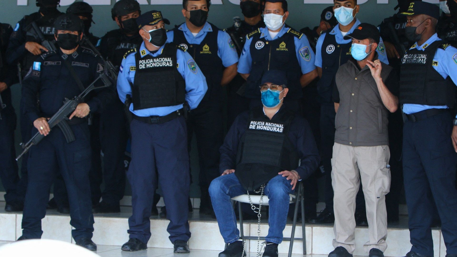 Former Honduran president Juan Orlando Hernandez sits, his ankles cuffed, surrounded by police in bulltproof vests and masks