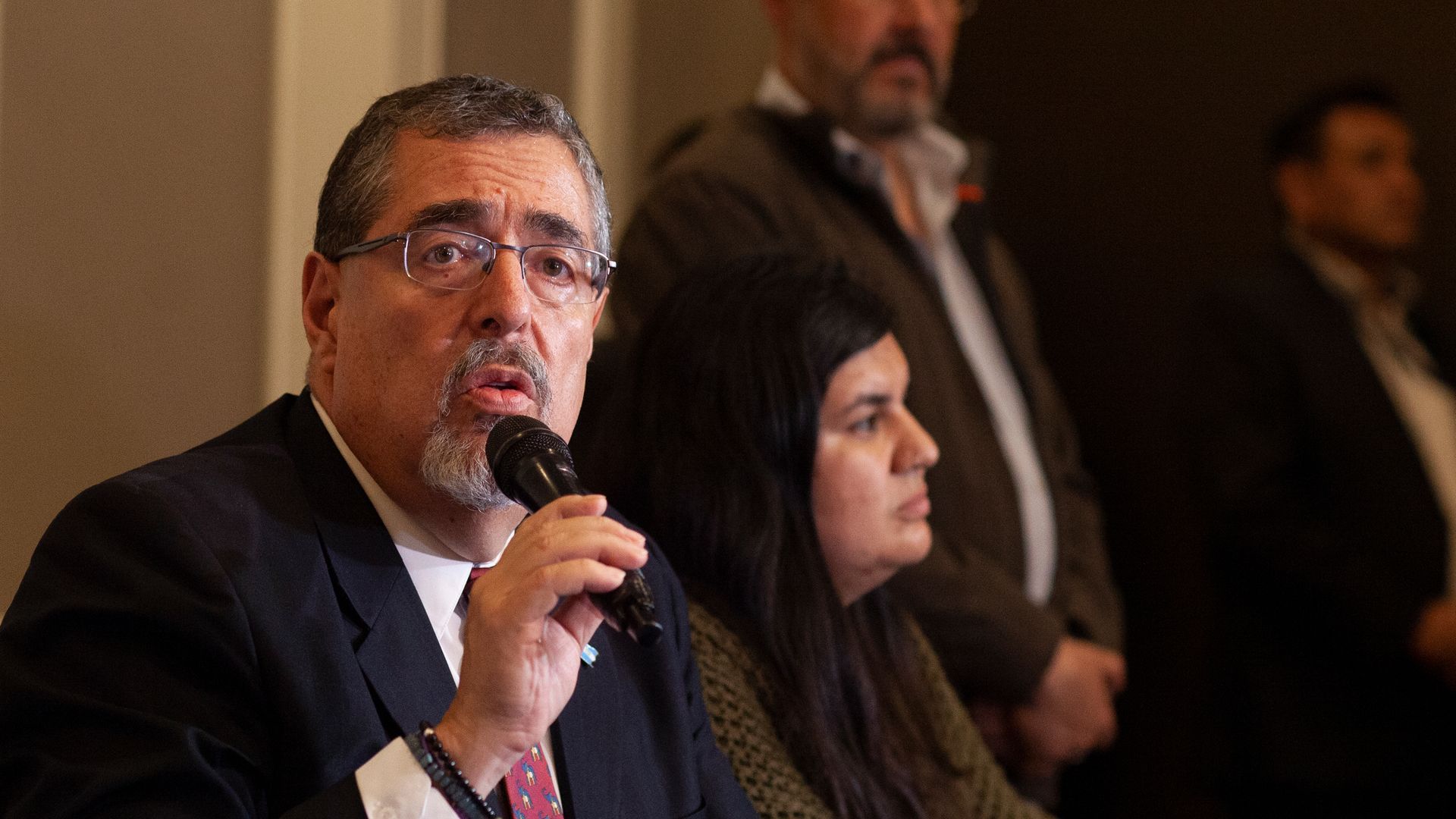 Guatemalan President-elect Bernardo Arevalo sits wearing a black suit and holding a black microphone to his face. he is wearing glasses and has a gray goatee.