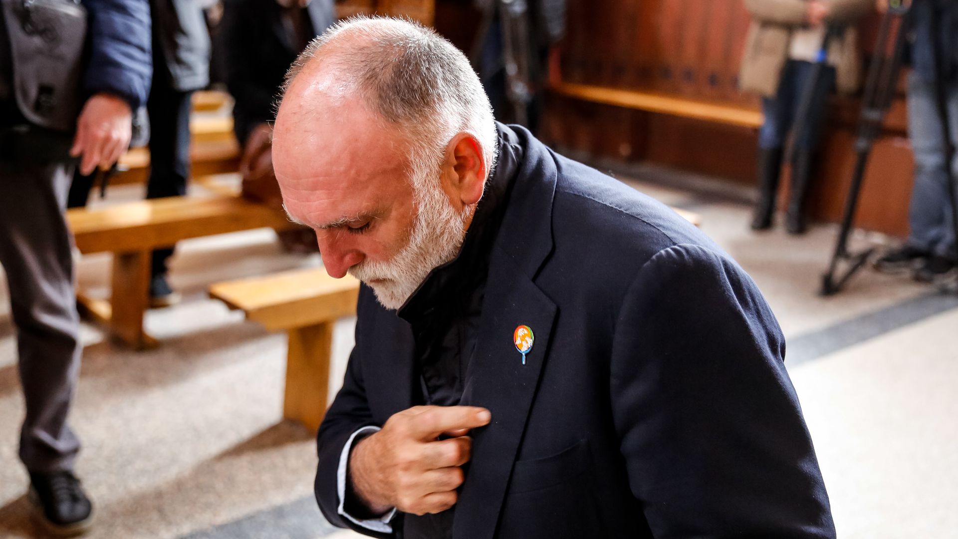 Jose Andres kneels, holding his hand over his heart, while wearing a World Central Kitchen pin on his jacket, at a memorial for a victim.