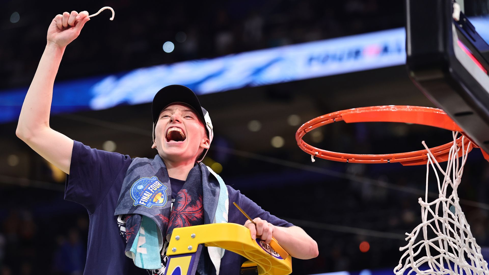 A basketball player climbs to the top of a ladder and holds a piece of white basketball net in her hand and celebrates