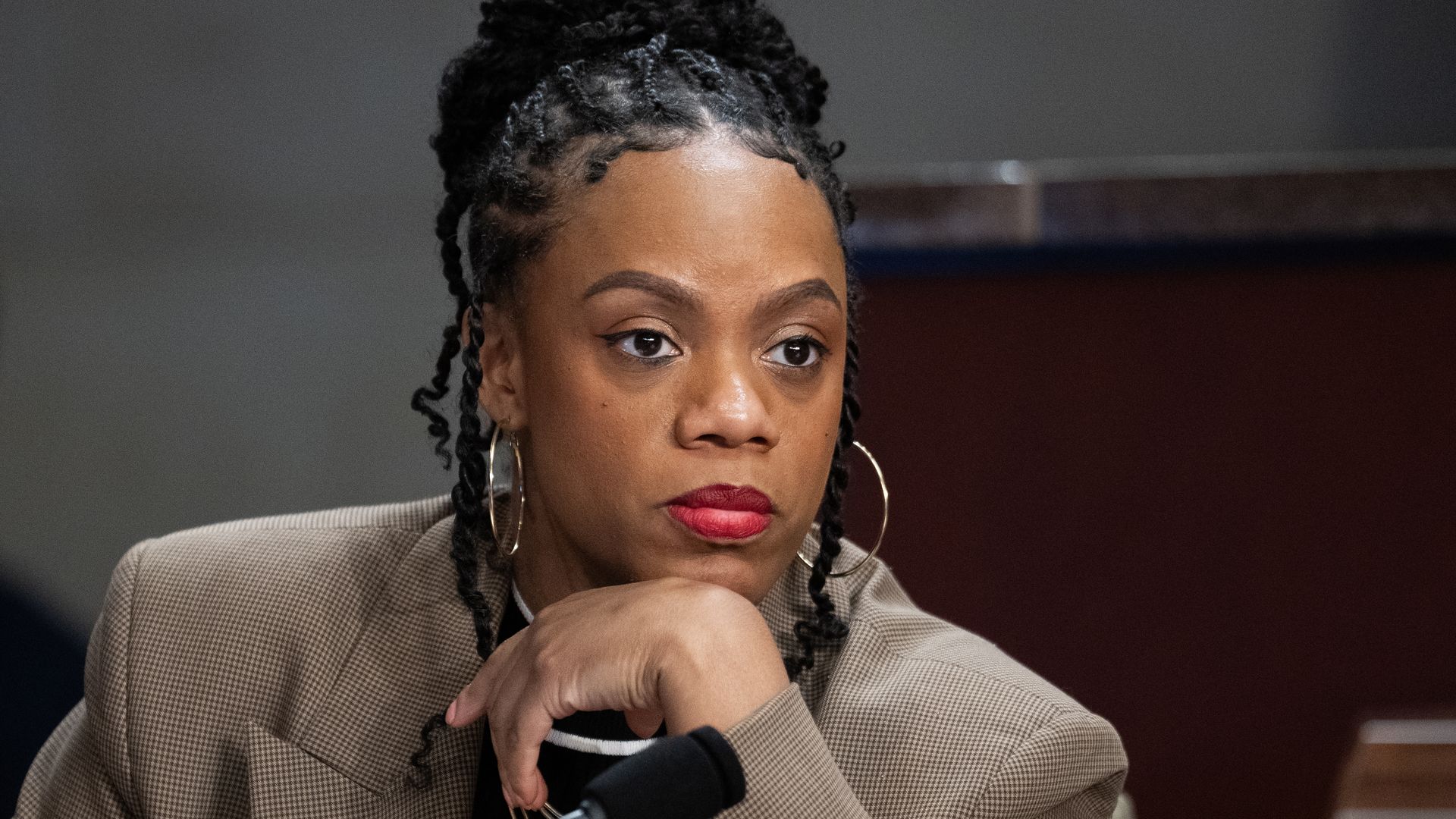 Woman with braided hair in a tan checkered blazer and red lipstick rests her chin on her hand, looking intently forward with large hoop earrings and a serious expression, at a microphone.