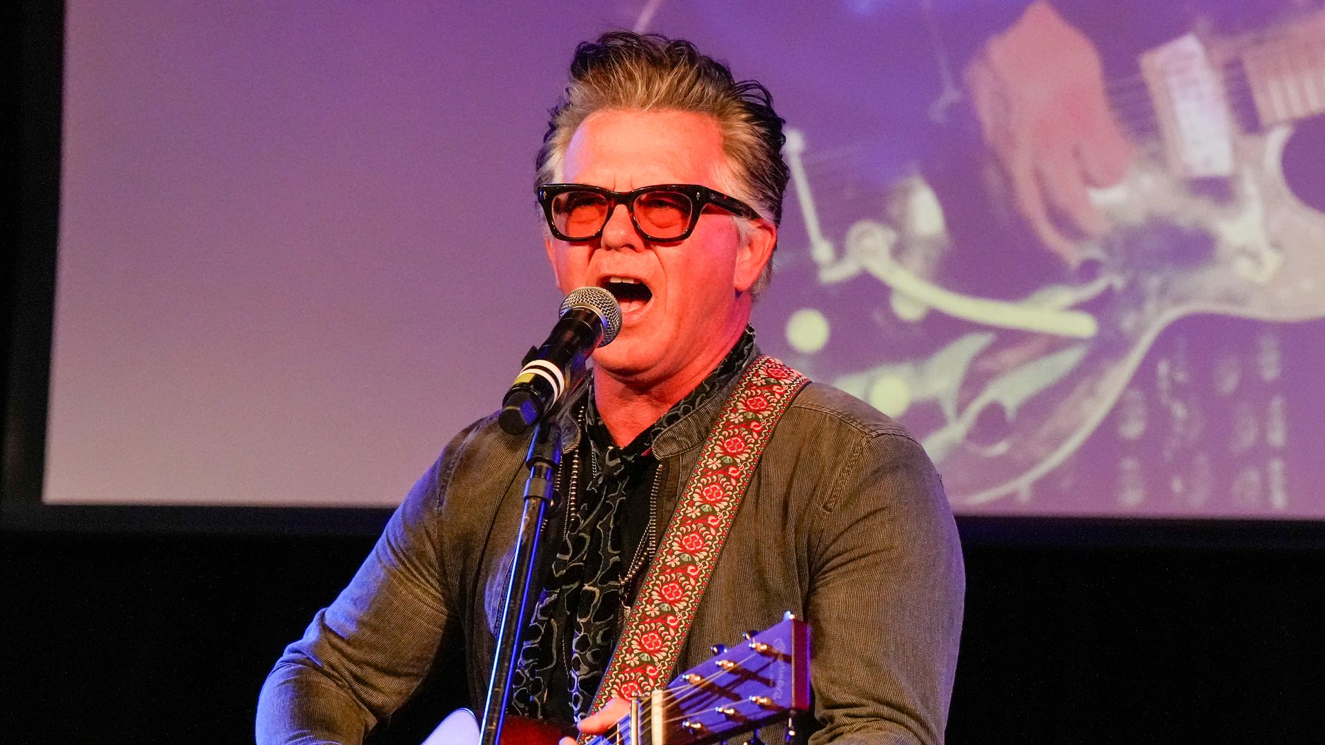 Man with gray-streaked hair and black glasses singing into a microphone, playing an acoustic guitar with a red patterned strap, against a purple backdrop with a projected image.