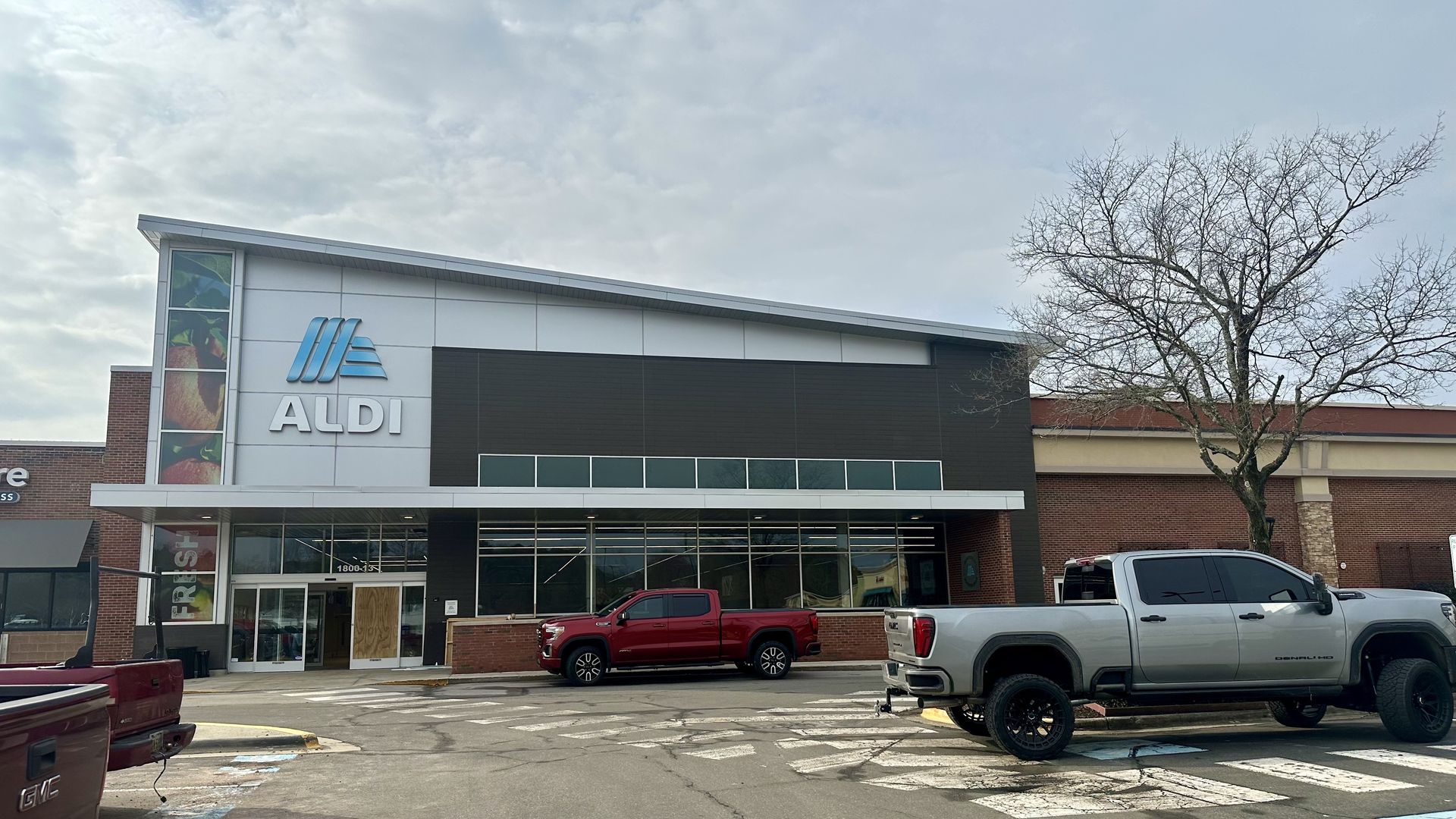 Exterior of an Aldi grocery store with a modern facade, black and white panels, and Aldi logo. Several pickup trucks parked in front under a cloudy sky and a leafless tree nearby.