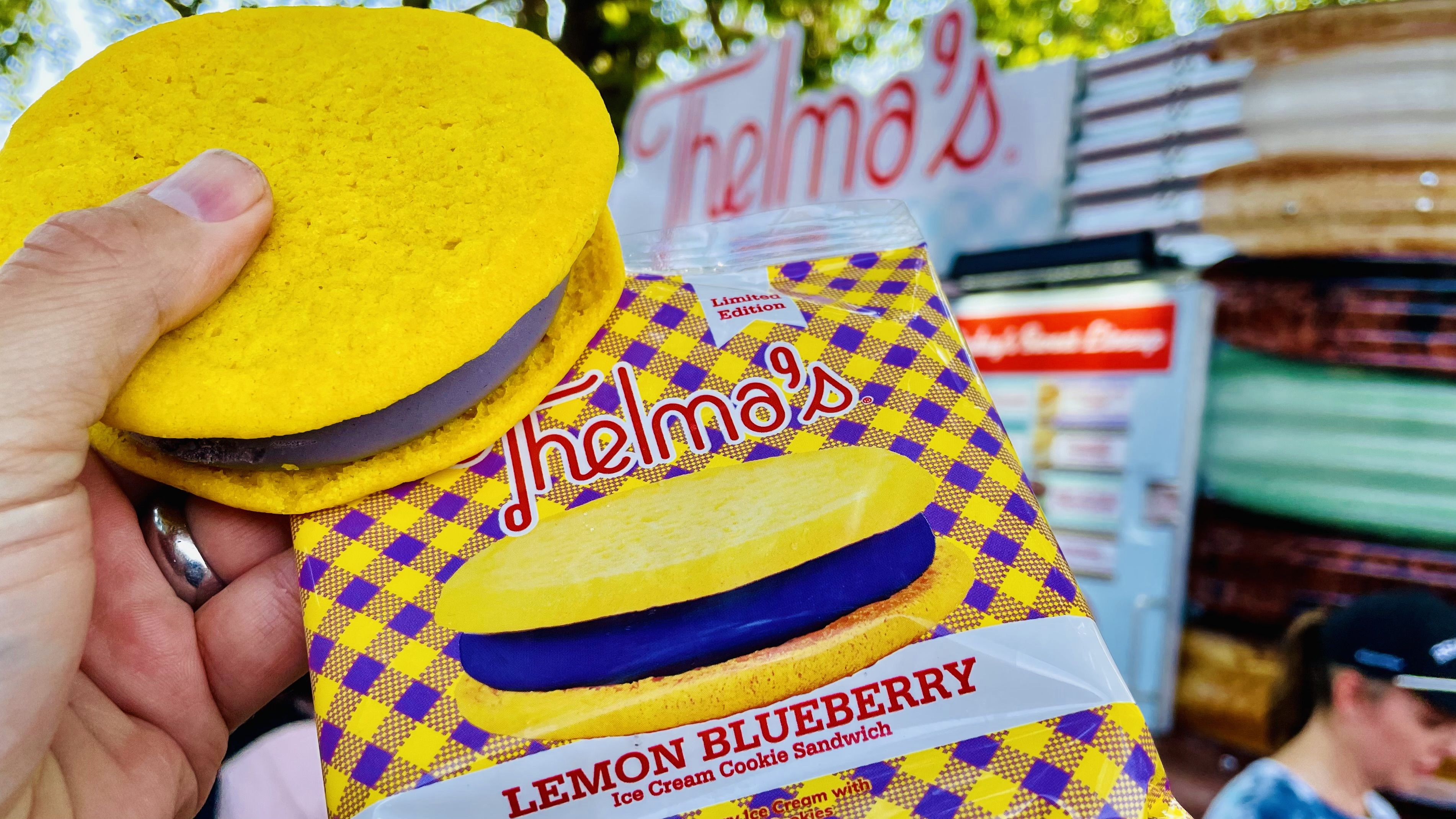 Hand holding a yellow lemon blueberry ice cream cookie sandwich beside its purple and yellow checkered Thelma's packaging outdoors with a blurred person and food truck in the background.