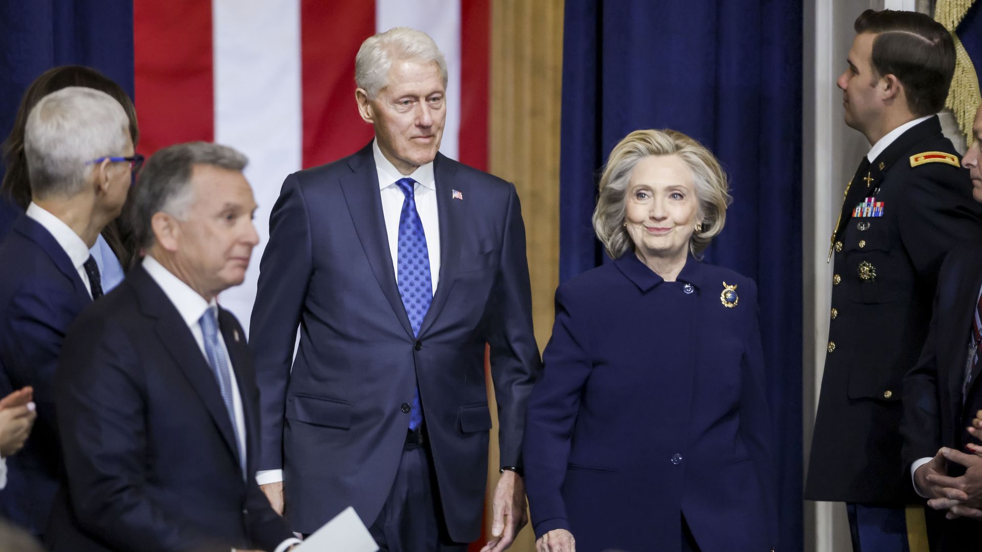 Bill and Hillary Clinton entering a formal event, with Bill in a dark suit and blue tie, and Hillary in a dark coat with a brooch, against a backdrop of American flag colors.