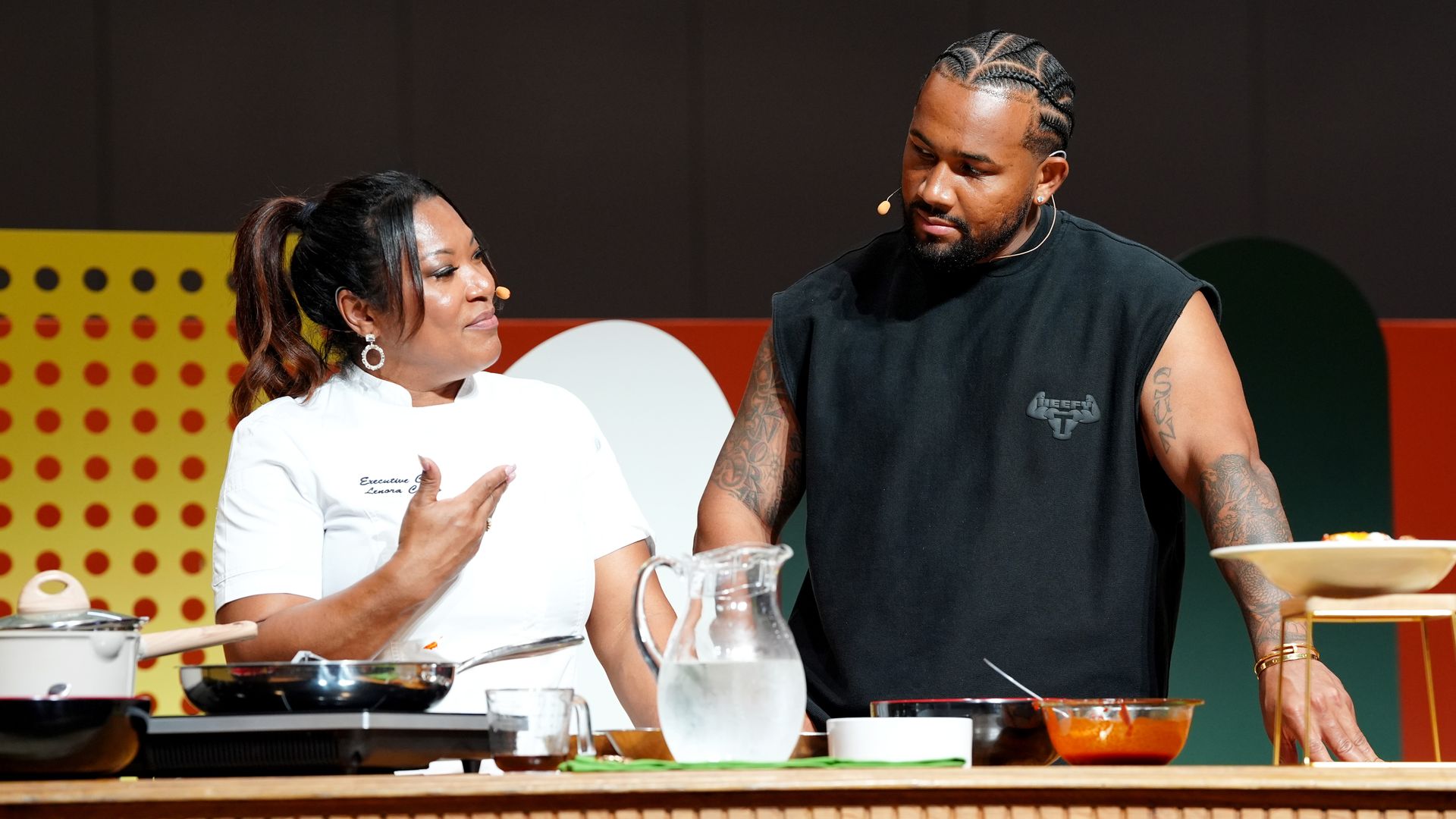A woman in a chef's coat speaks with a man in a black shirt as they stand behind the counter of an onstage demo kitchen.