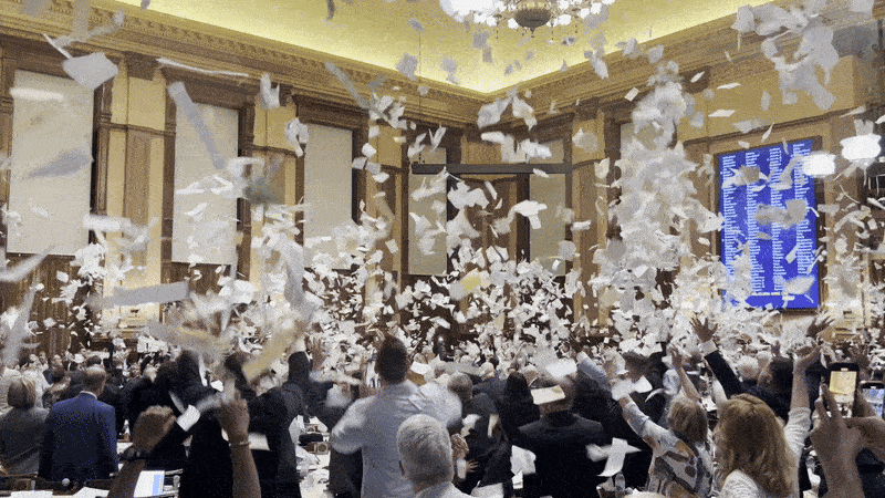 A GIF of people throwing papers into the air in a large legislative chamber
