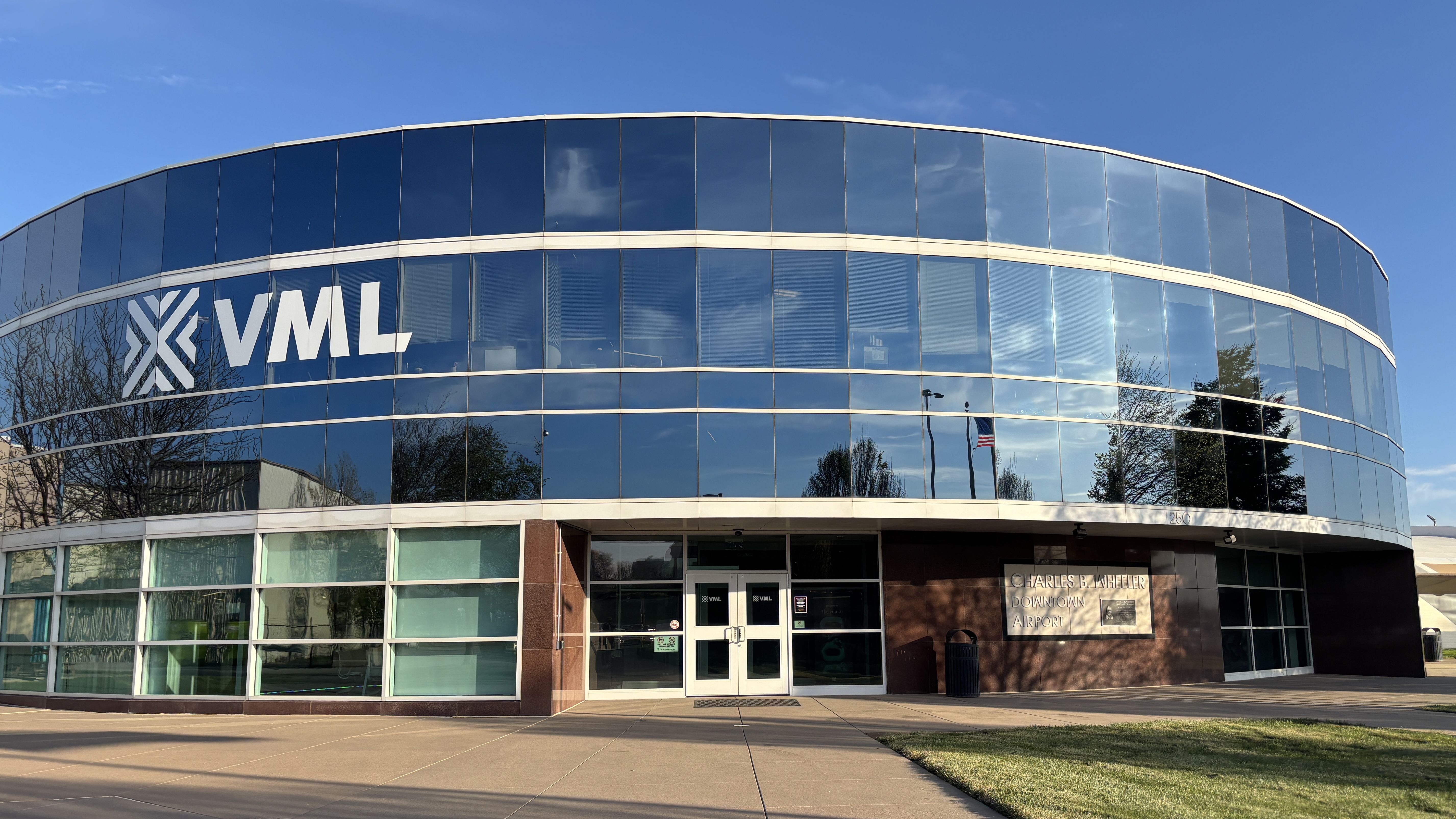Curved glass building with blue-tinted panels and white bands. Large 'VML' logo on the left; glass entrance at center; reflections of trees and a flag. Brick wall sign reads "Charles B. Wheeler Downtown Airport".