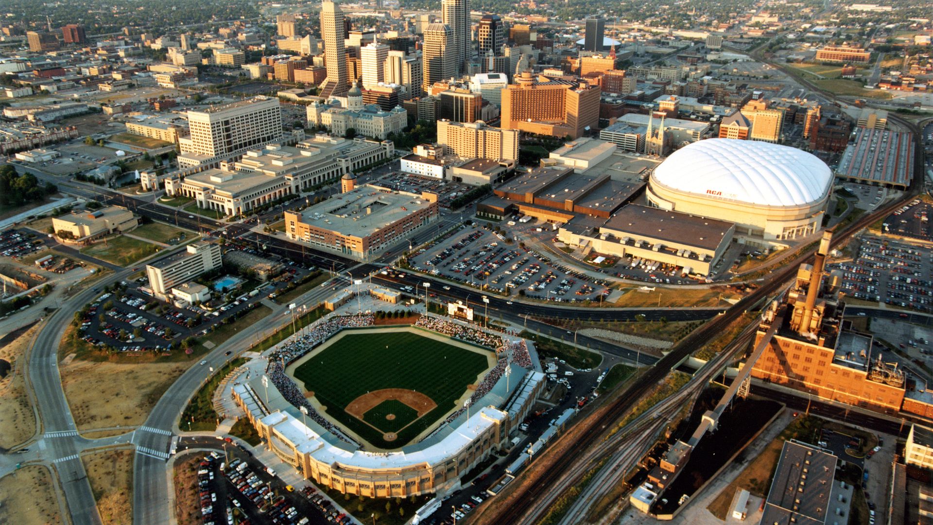 An aerial view of a downtown skyline with tall skyscrapers, a large white-domed arena, surrounding low-rise buildings, a baseball field in the foreground, and rail lines and roads at sunset.