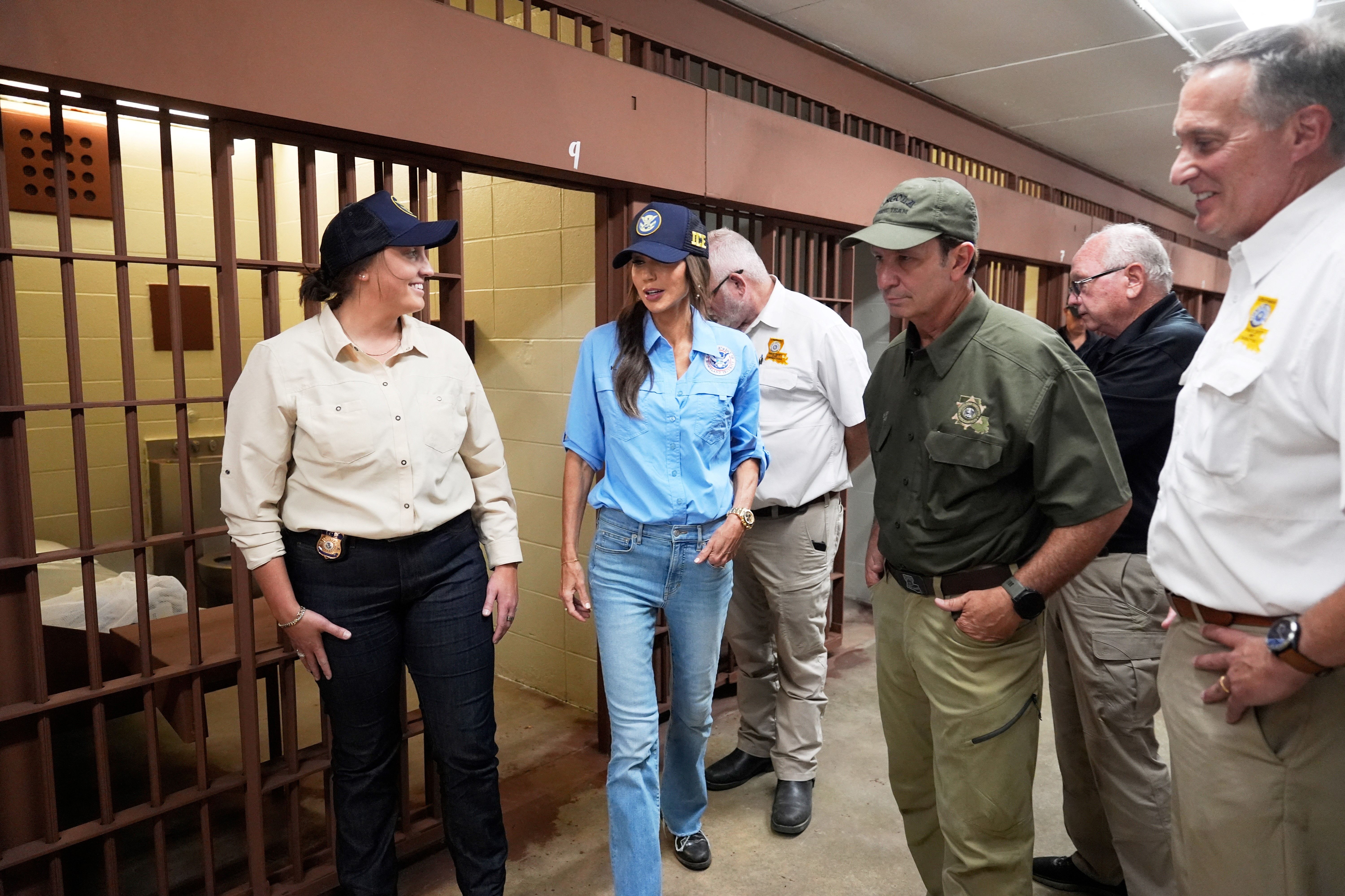 Five people in casual but professional attire stand inside a jail hallway near metal-barred cells, engaged in conversation. The walls are beige, and the floor is concrete.