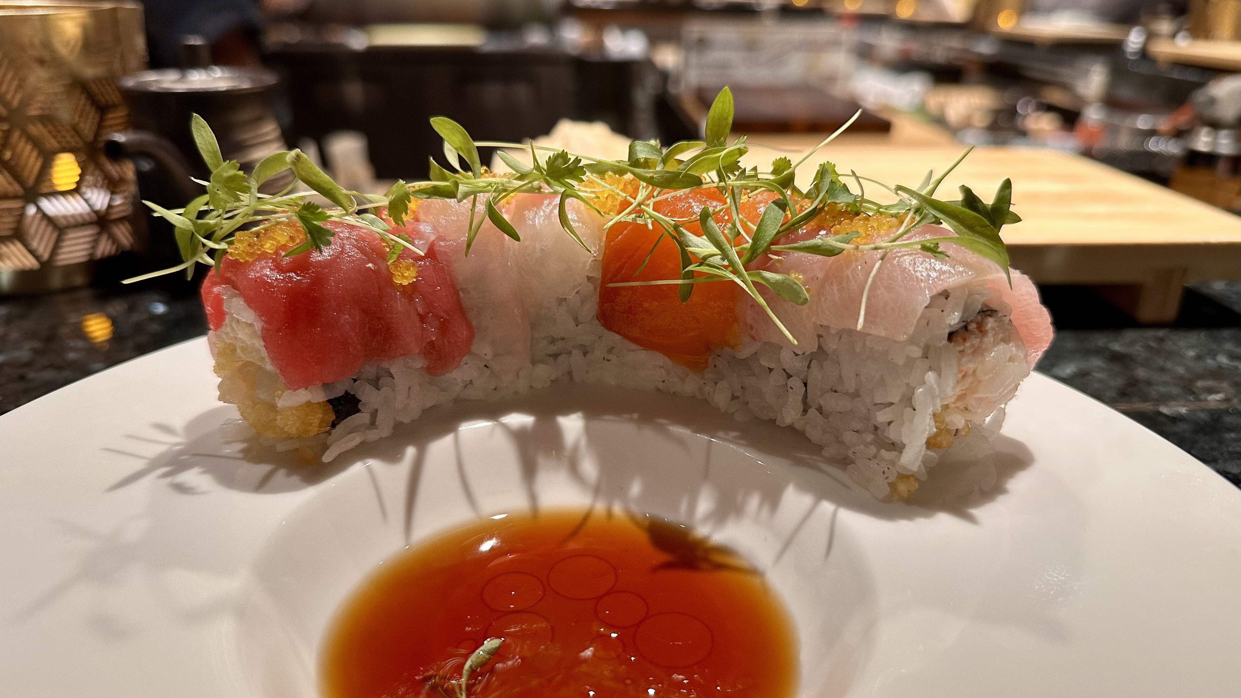 Close-up of colorful sushi rolls topped with green herbs and orange roe, served on a white plate with a pool of soy sauce in the center, set against a blurred restaurant background.
