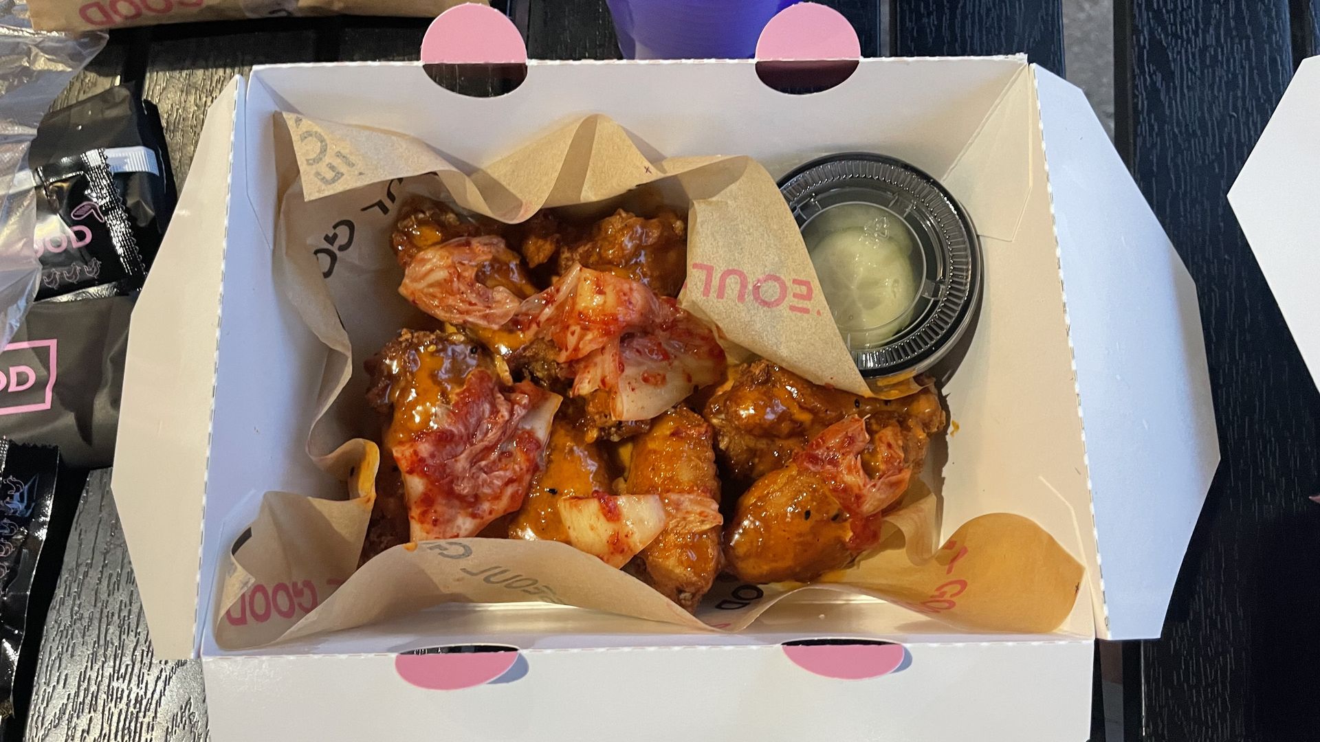 A paper to-go box is filled with chicken wings covered in kimchi.