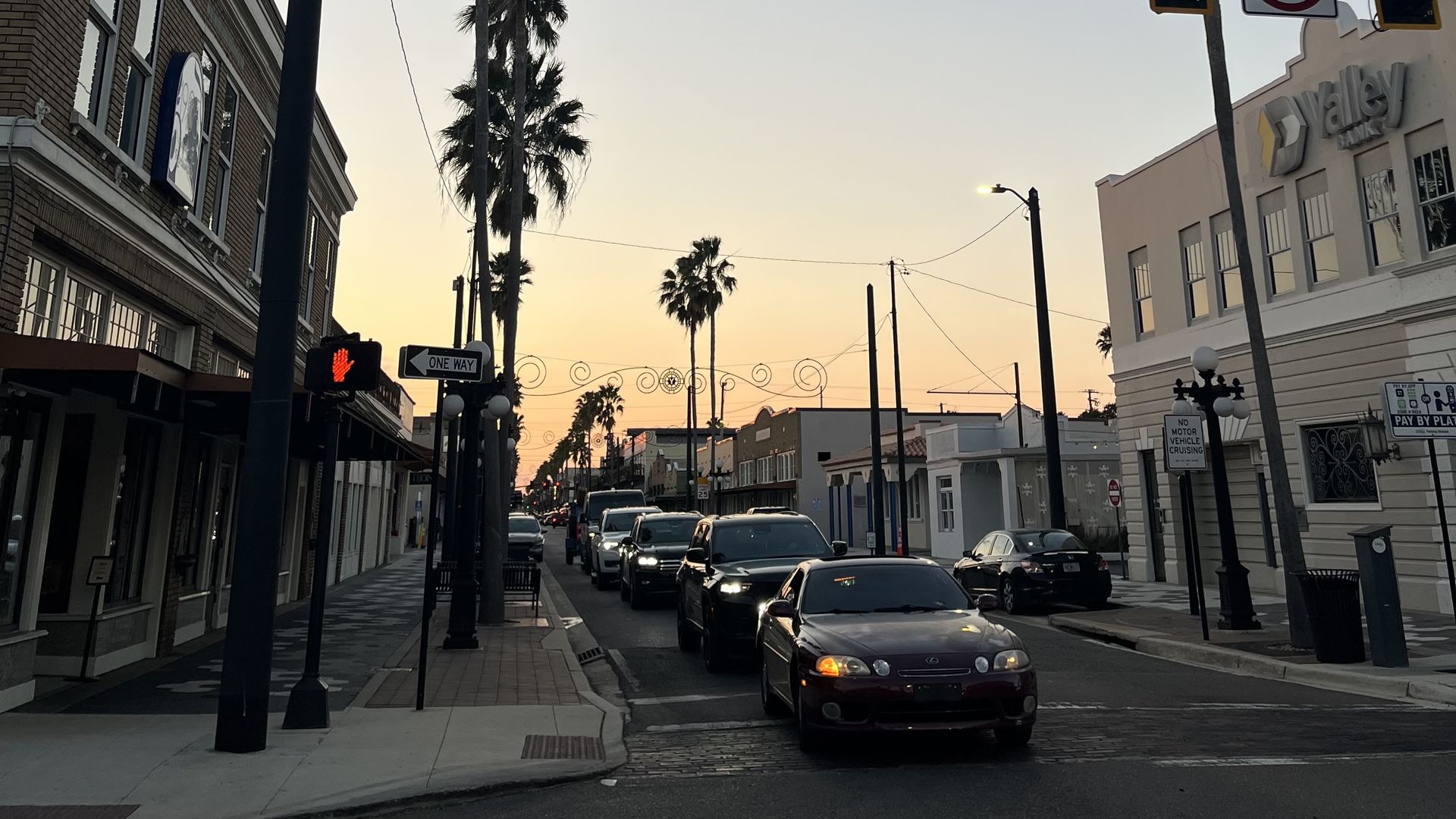 Sunset street scene with palm trees lining a one-way road filled with cars stopped at a red pedestrian signal, flanked by buildings including a bank on the right side.