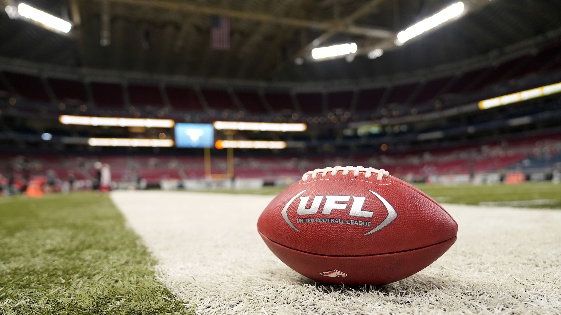 Brown UFL (United Football League) football on white sideline of a green football field inside a stadium with empty red seats in background.