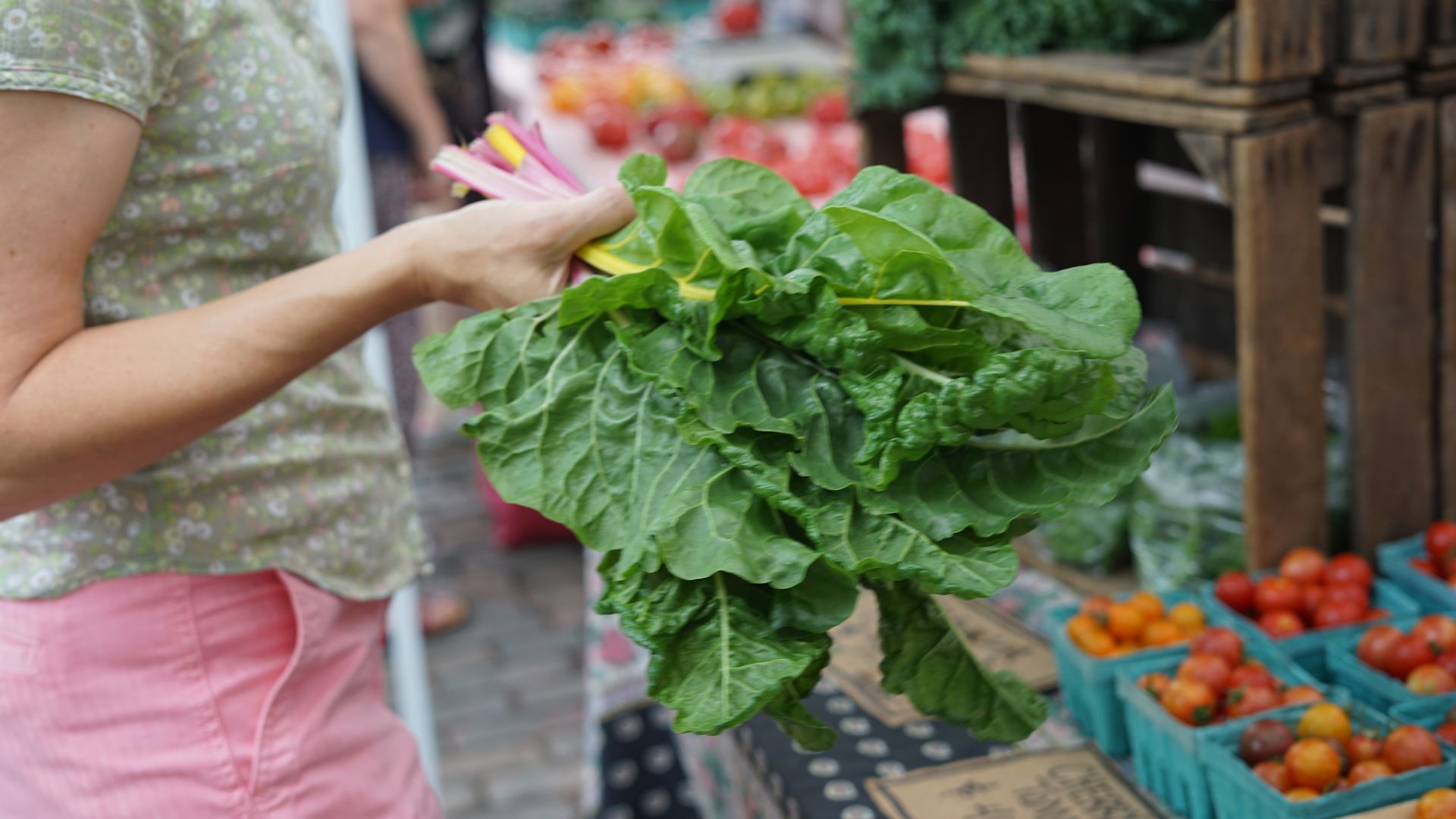 Person at an outdoor market holds a large bunch of green leafy vegetables with pink stems, wearing a green floral shirt and pink shorts; blurred crates of produce in the background.