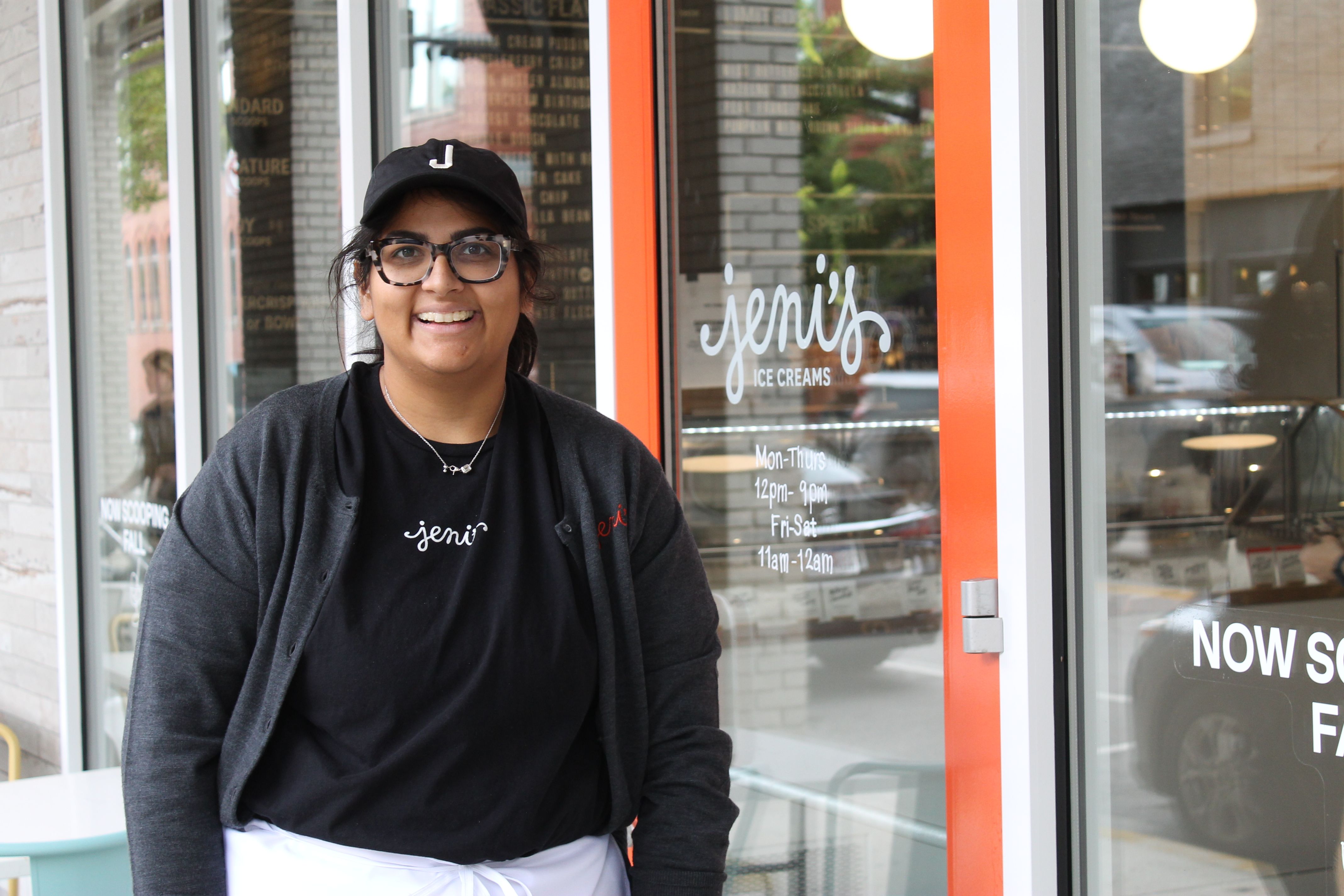 Smiling person wearing a black cap with a white J, glasses, and a dark shirt, standing outside the storefront of "jeni's Ice Creams" with orange trim.
