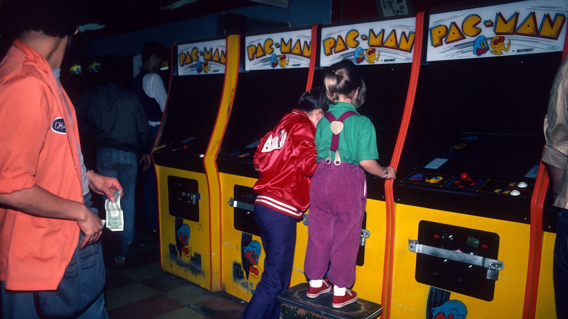 Two young children playing Pac-Man in an arcade. One stands on a stool to see the screen.