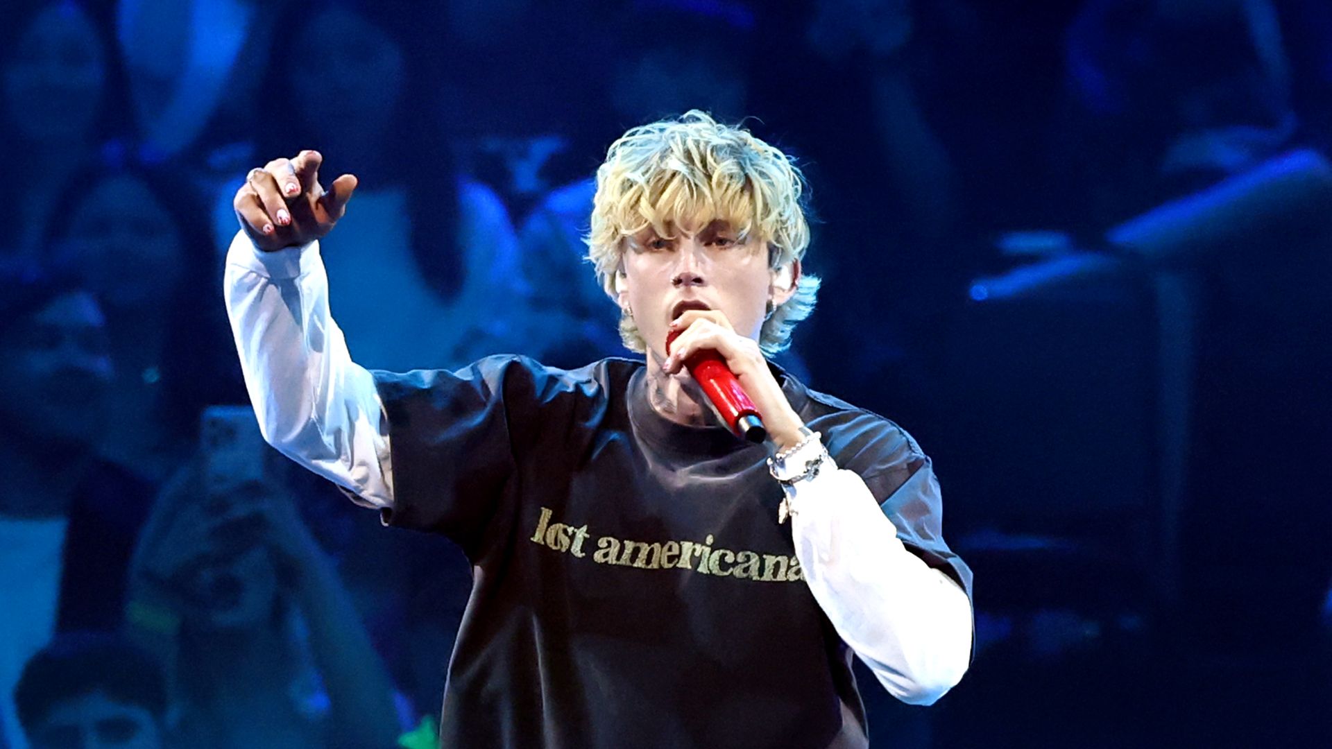 Blond male singer with curly hair performs onstage holding a red microphone, wearing a black shirt with white long sleeves that says "lost americana". Audience blurred in background.
