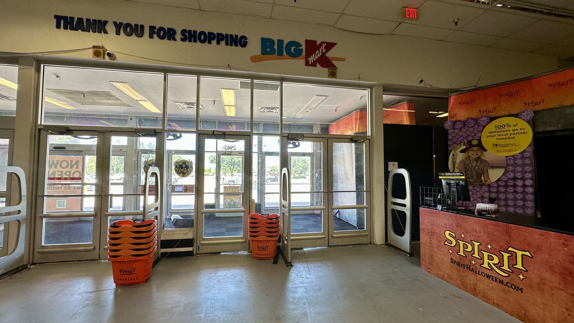 Store entrance with glass doors, a large THANK YOU FOR SHOPPING sign and a blue BIG Kmart logo. Orange baskets by the door; Spirit Halloween display on the right with an orange banner and a yellow donation circle.