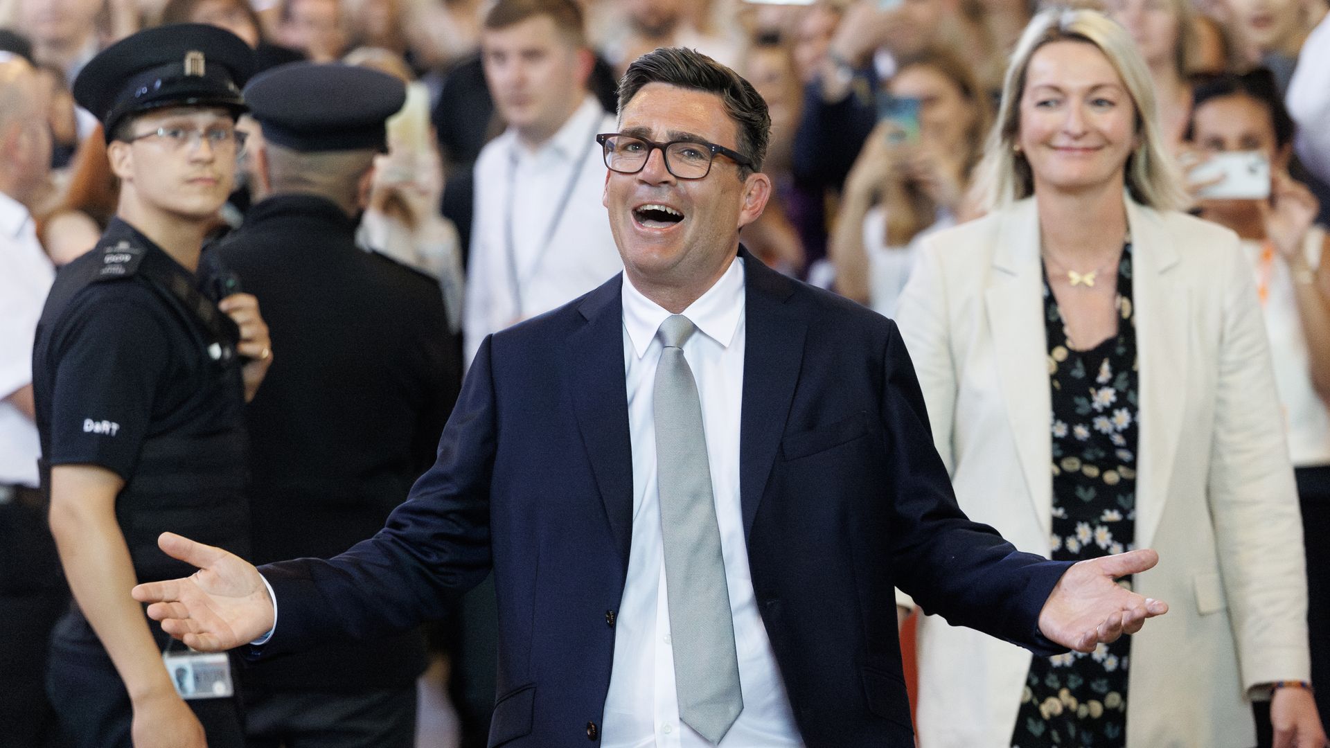 UK Labour MP Andy Burnham, in a navy suit and gray tie with glasses smiles, arms open, walking through a crowd; a security guard on the left and a woman in a light blazer smiles on the right.