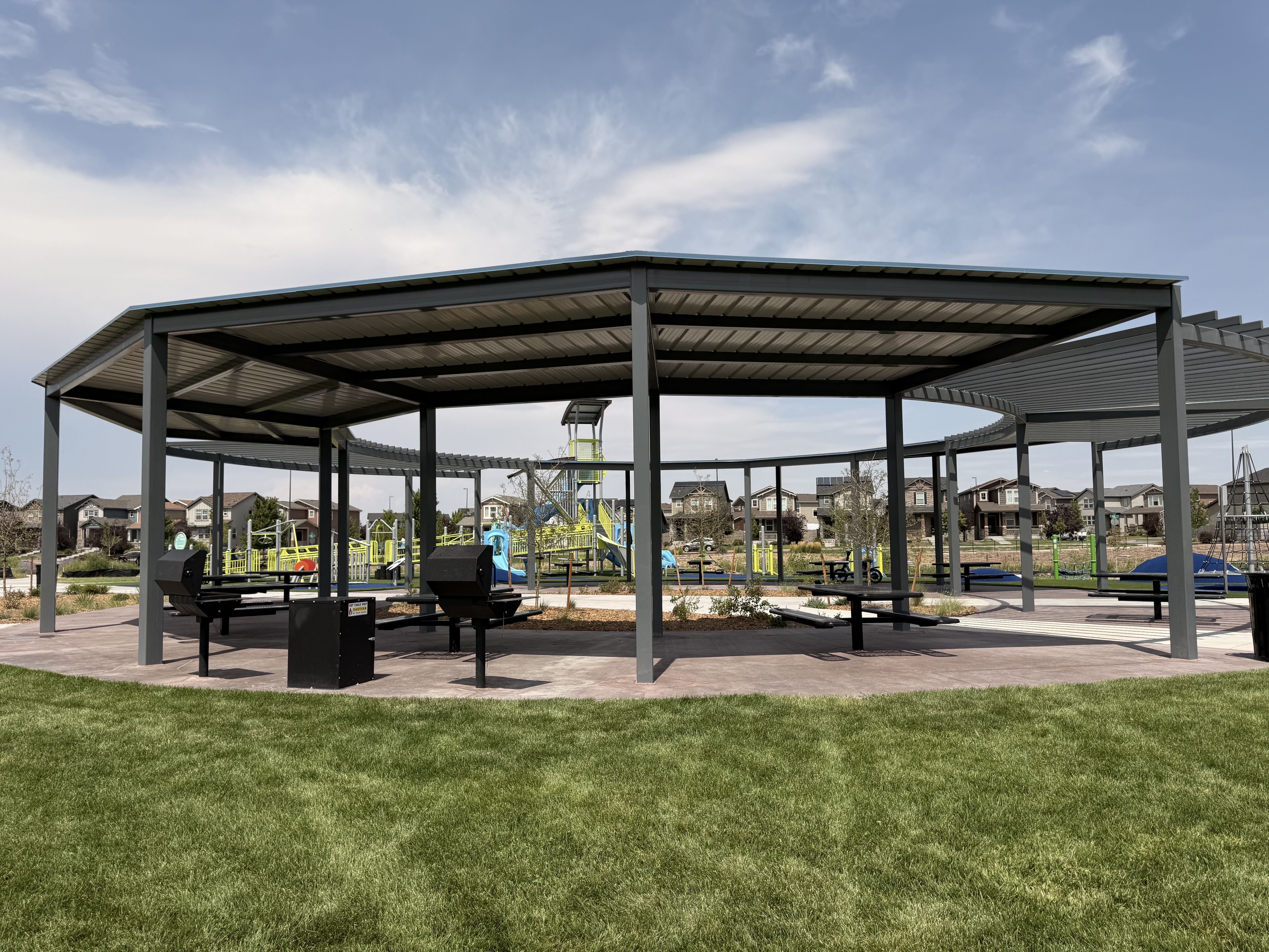 A large gray metal pavilion with open sides and built-in grills sits over concrete; a green lawn in the foreground, a playground and houses in the background under a blue sky.