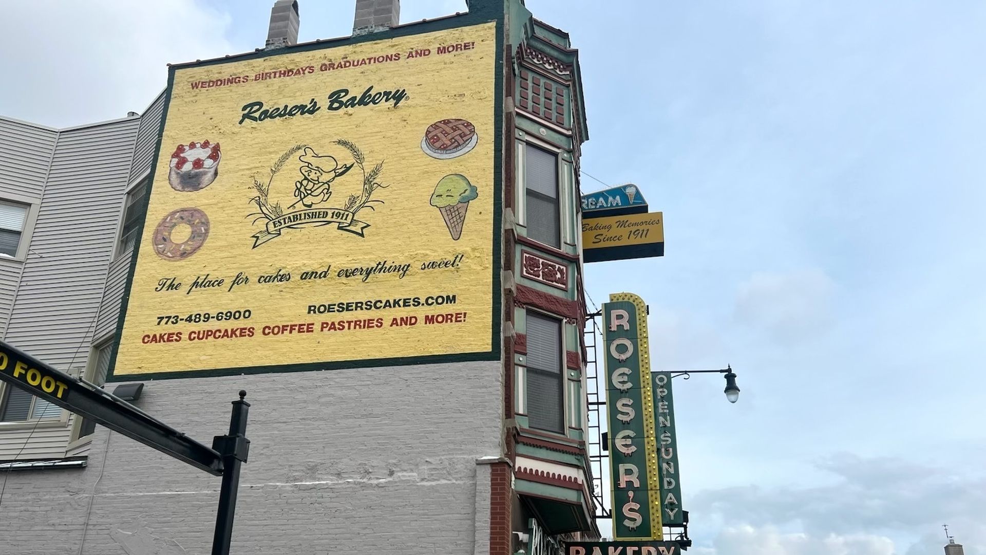 Yellow painted sign on a brick wall. The sign has different desserts painted on it. The building has a neon vertical Roeser's sign.