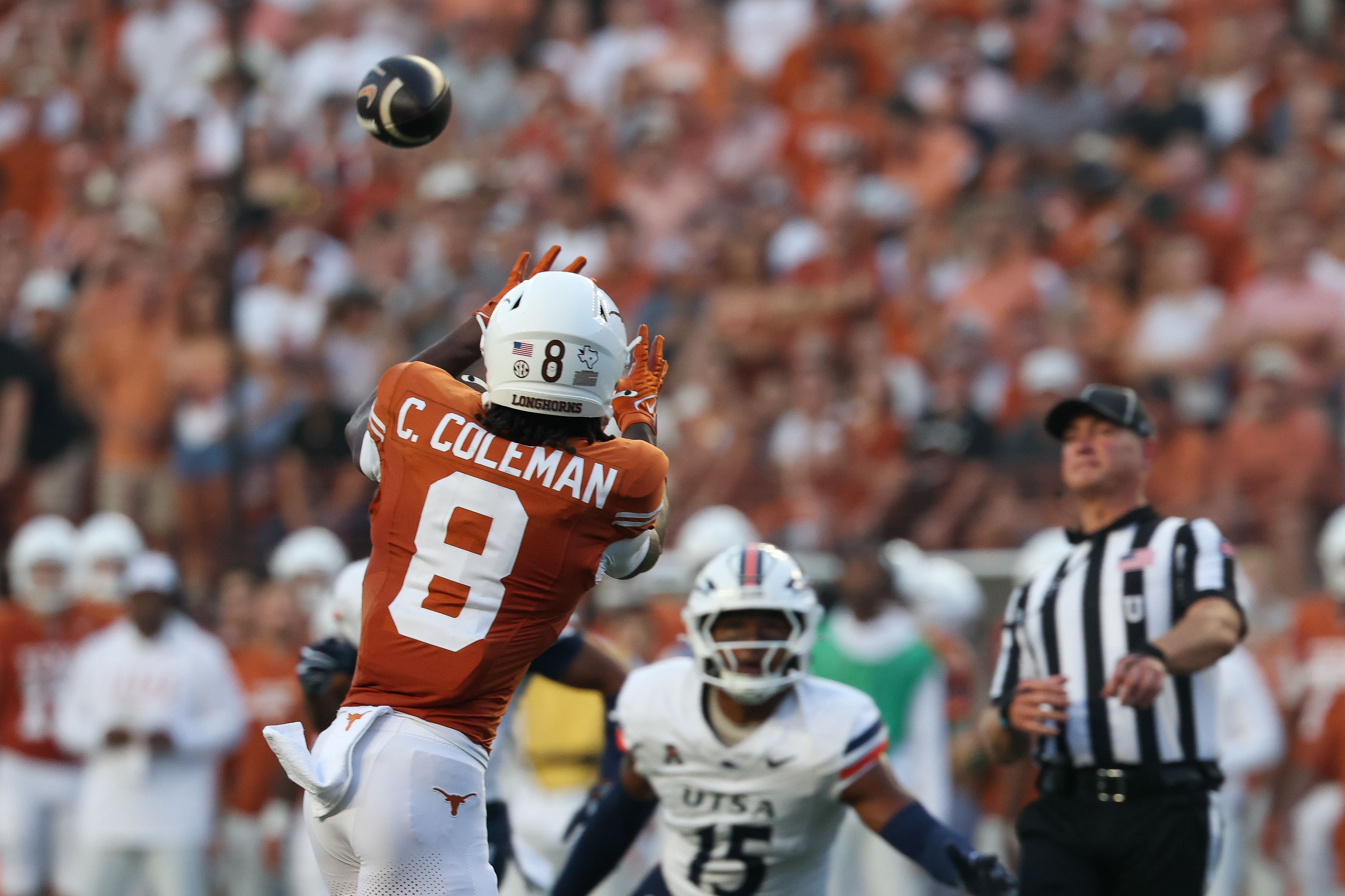 Texas Longhorns football player in orange jersey, C. Coleman, #8, reaches up as a football sails overhead; blurred crowd and a referee in black-and-white behind.