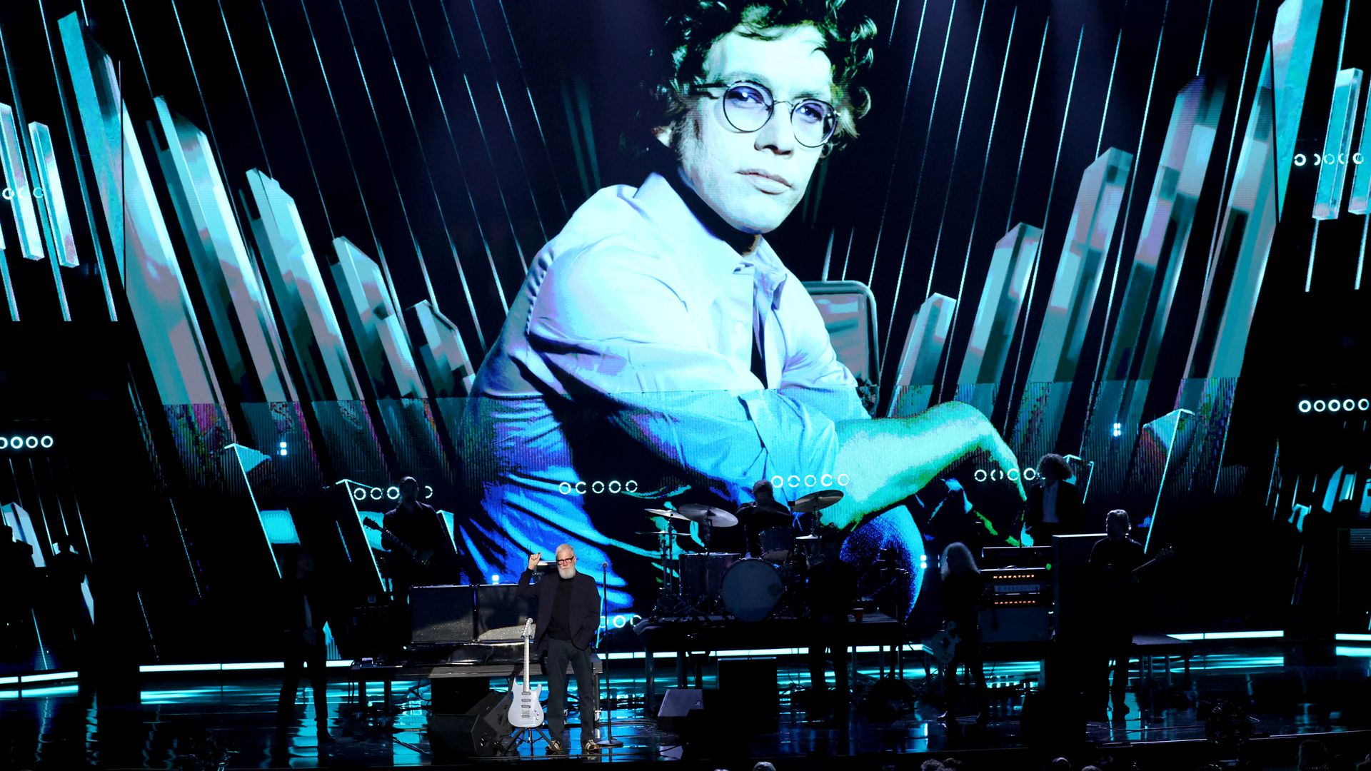 Man with white beard and glasses stands with guitar on stage in dim light, large blue-toned portrait of young man wearing glasses displayed on screen behind band setup with drums and guitars.