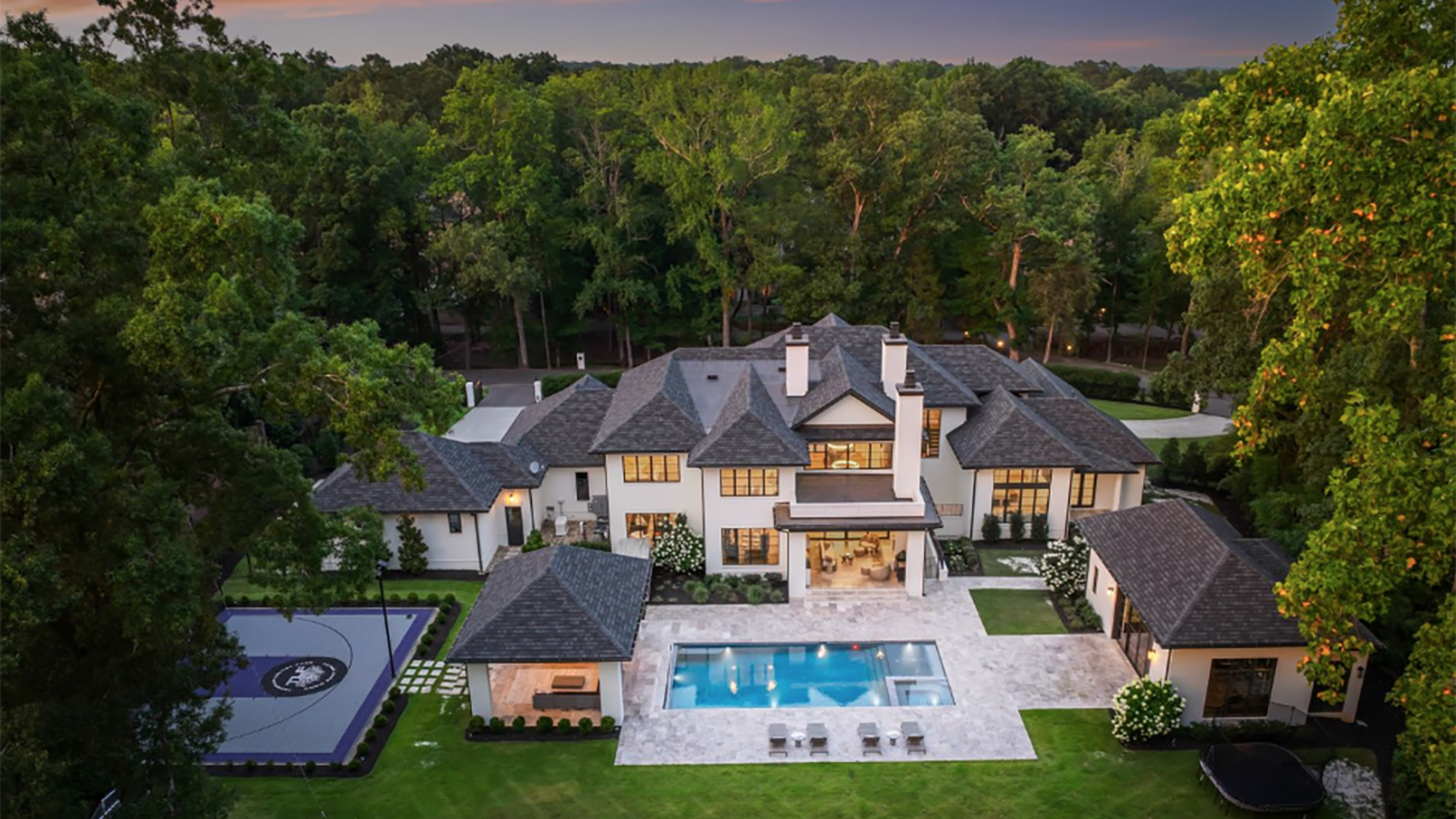 A large luxury house with gray gabled roofs, light walls, a swimming pool, outdoor seating, a covered patio, a small building, a basketball court, and surrounding green trees at sunset.