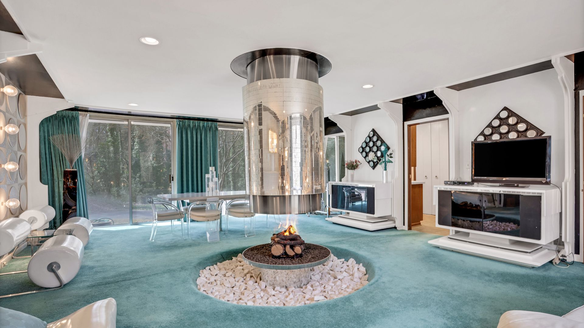 A mid-century living room with aqua shag carpet, white ceiling, and cylindrical hanging fireplace with mirrored tiles