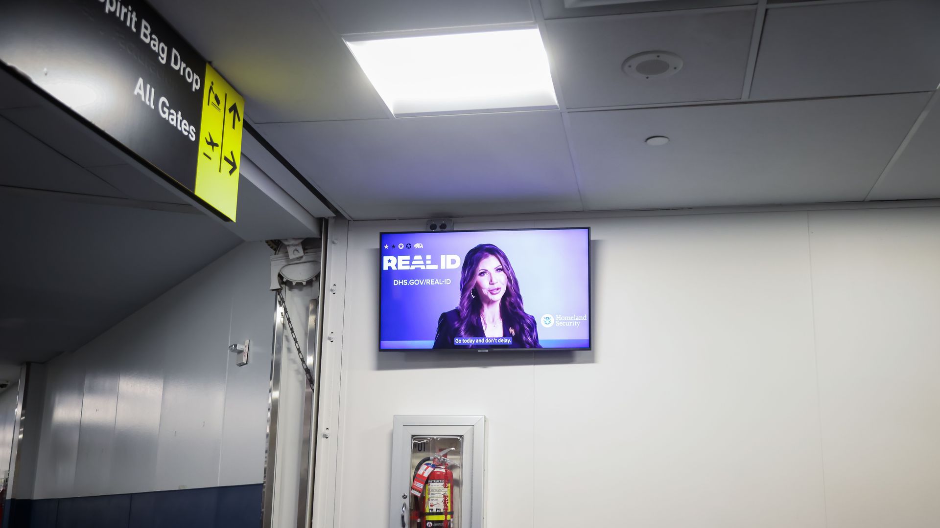 Airport interior with a wall-mounted TV showing a Homeland Security ad about REAL ID, a fire extinguisher in a case below, and a yellow Spirit Airlines sign indicating bag drop and all gates.