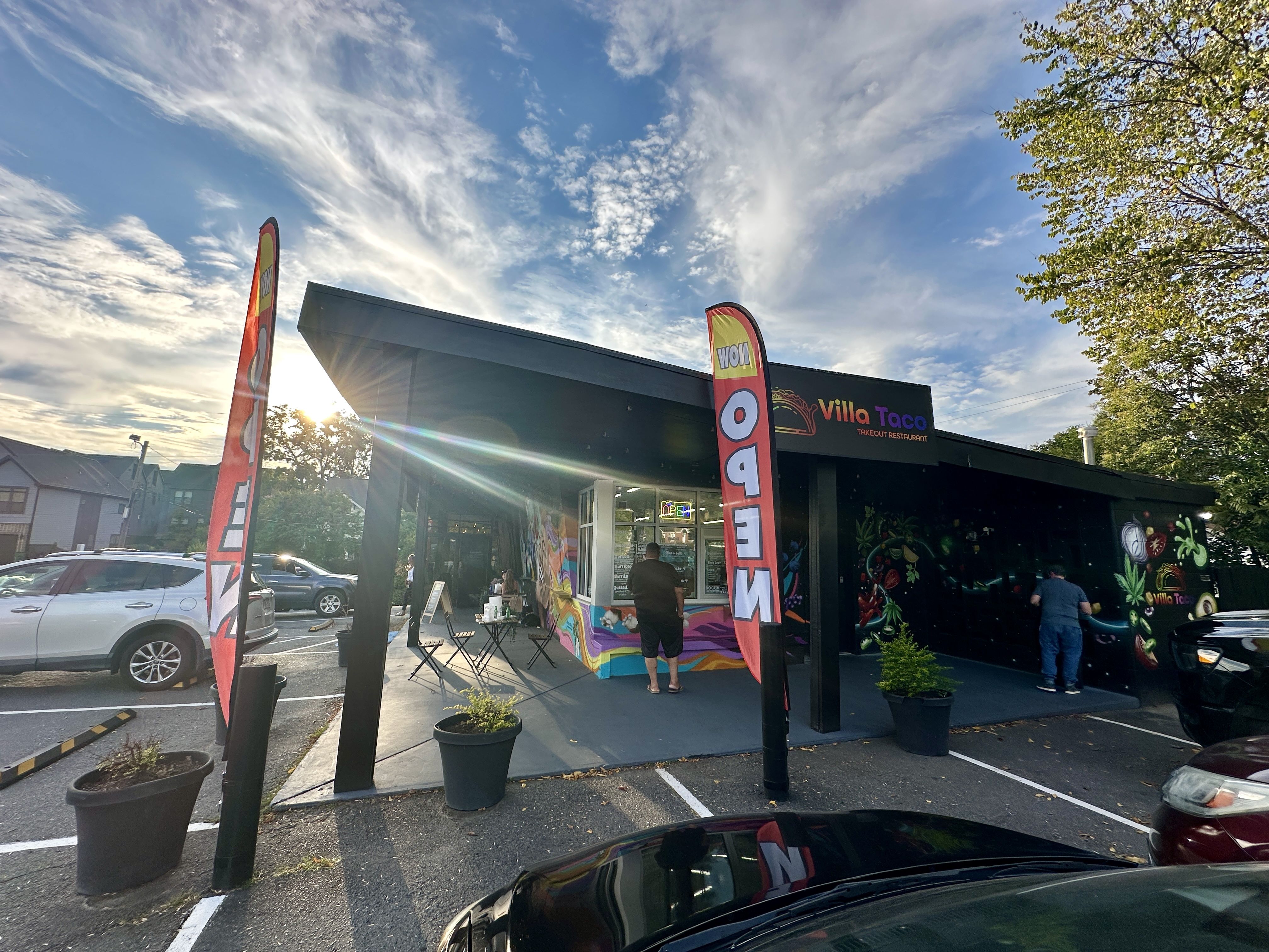 Villa Taco takeout restaurant with colorful mural, two vertical red "OPEN" flags, outdoor seating, and cars parked under a partly cloudy blue sky at sunset.