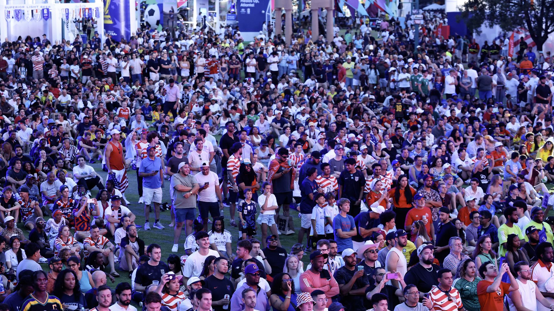 A large crowd of people wearing red and white striped shirts and other U.S.-affiliated colors watch a soccer game in a large public park.
