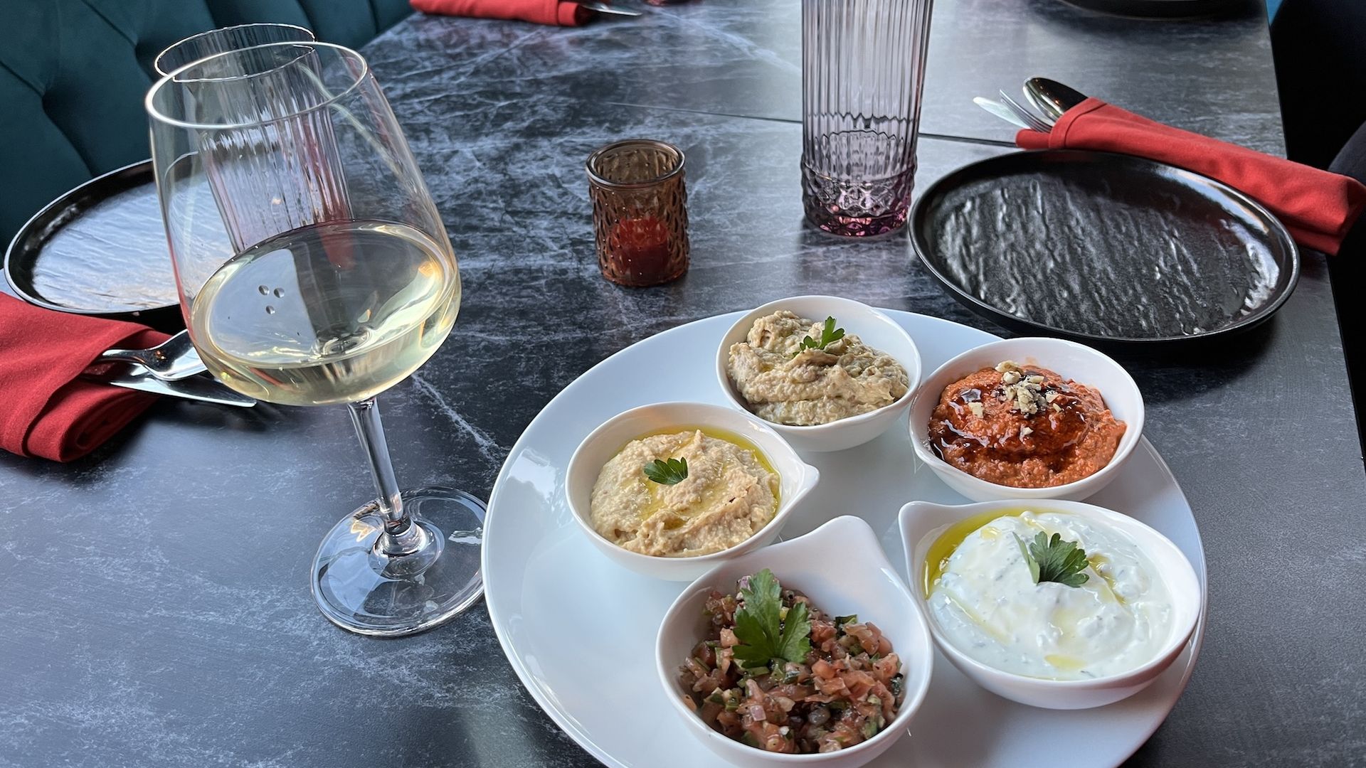 A white plate with five bowls of assorted dips including creamy, red, and herb-based varieties, garnished with parsley, next to a glass of white wine on a gray marble table.