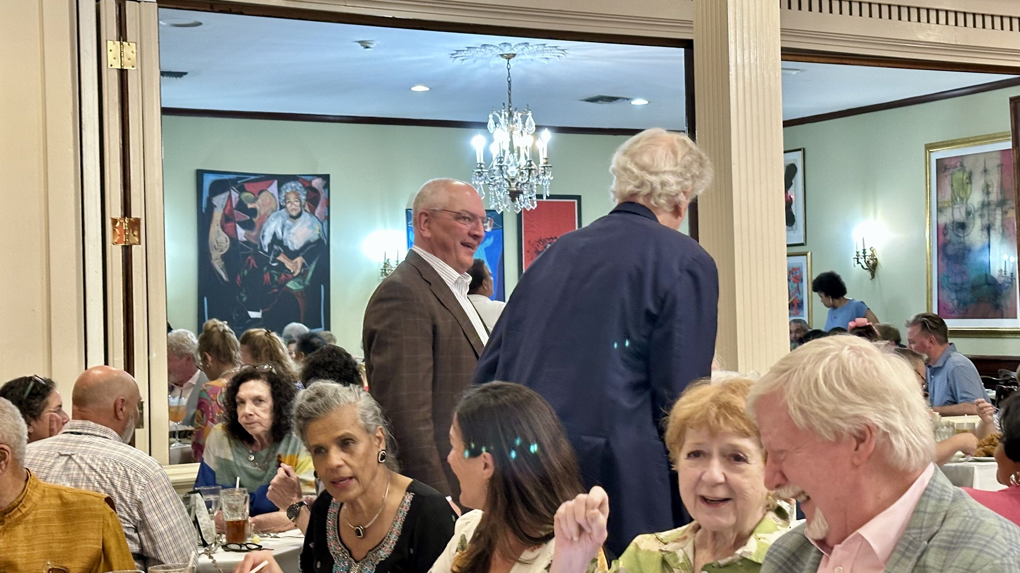 Crowded restaurant dining room with guests at tables. A man in a brown check blazer smiles at the camera; another man in a dark blue jacket stands with his back turned. An ornate chandelier and framed art on pale green walls.