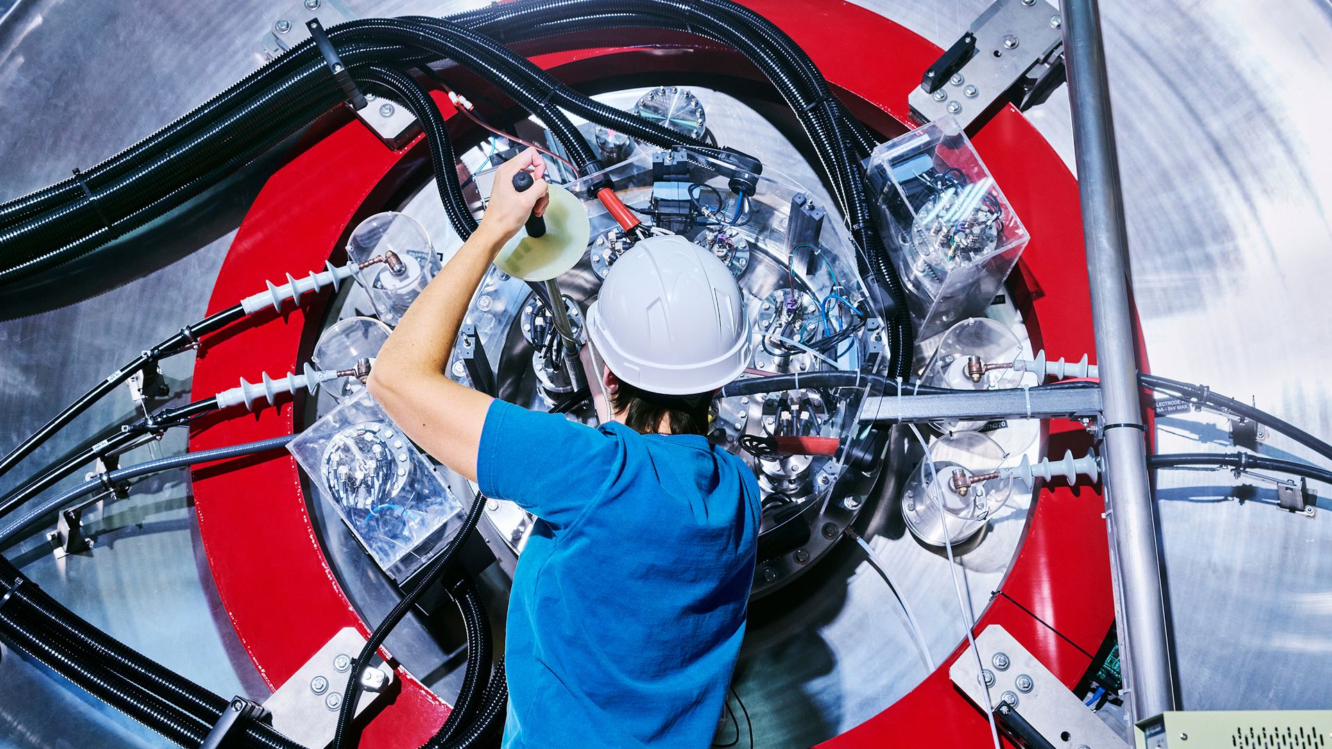 A person in a blue T-shirt and white hardhat holds a tool against a large, metal cylindrical machine with a red ring like a target.