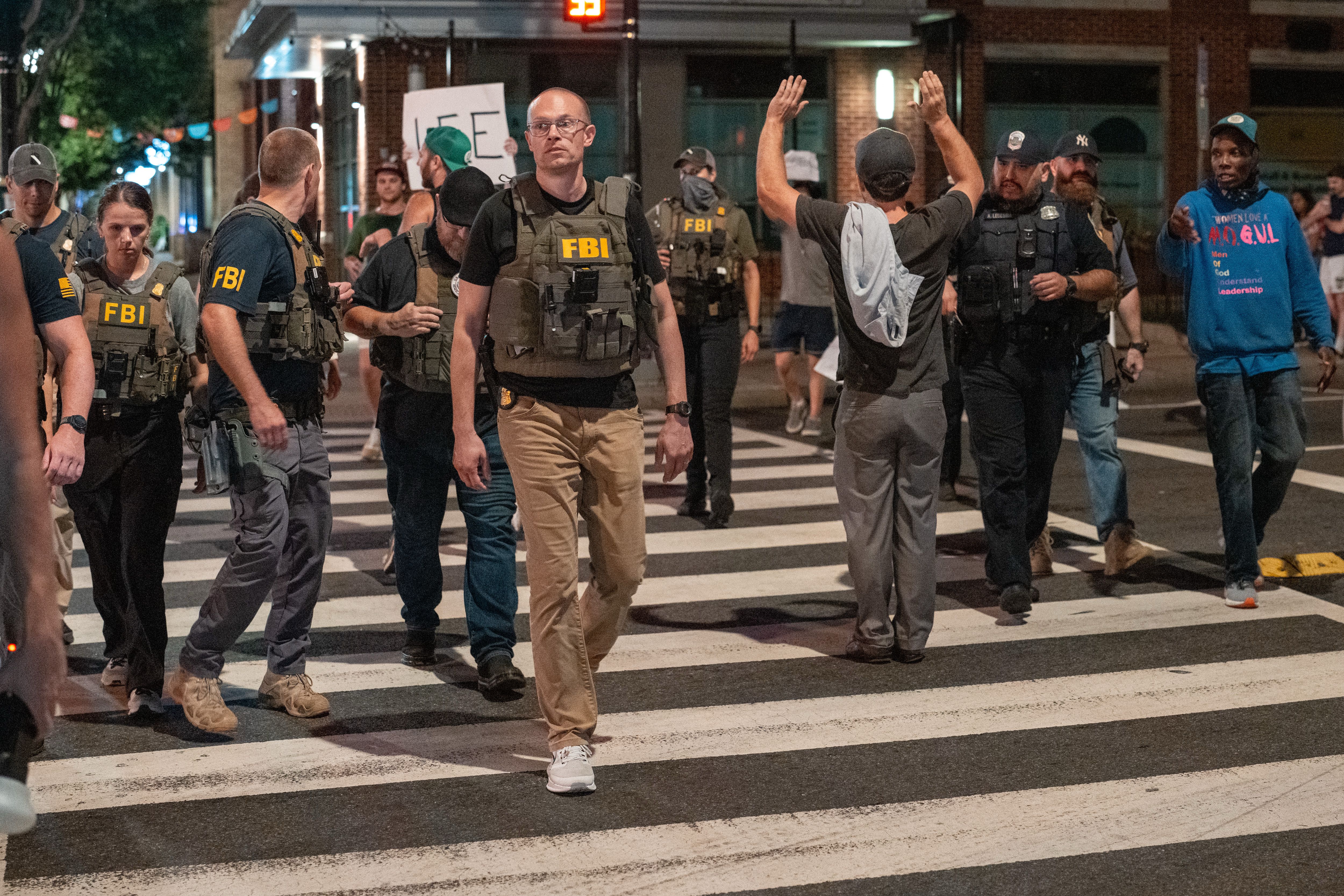 FBI agents and police officers wearing tactical gear face pedestrians, including one with hands raised, on a city street at night during a tense scene.