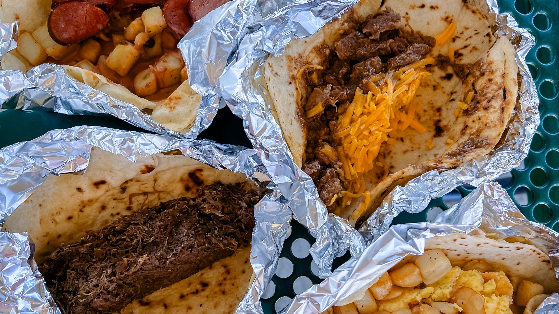Four tacos wrapped in foil, holding a variety of proteins and toppings. 