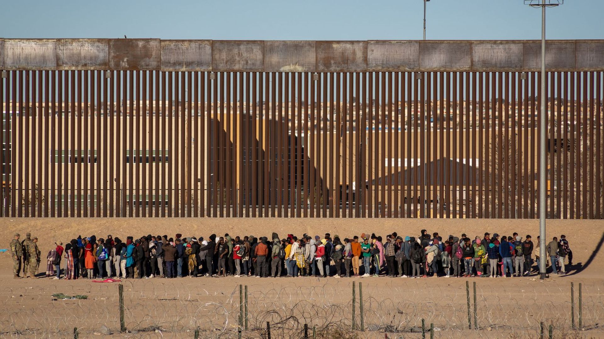 From a distance, dozens of migrants are shown line up in front of a tall border fence. They are in the desert, atop of sand, and there are several border patrol agents around them. 
