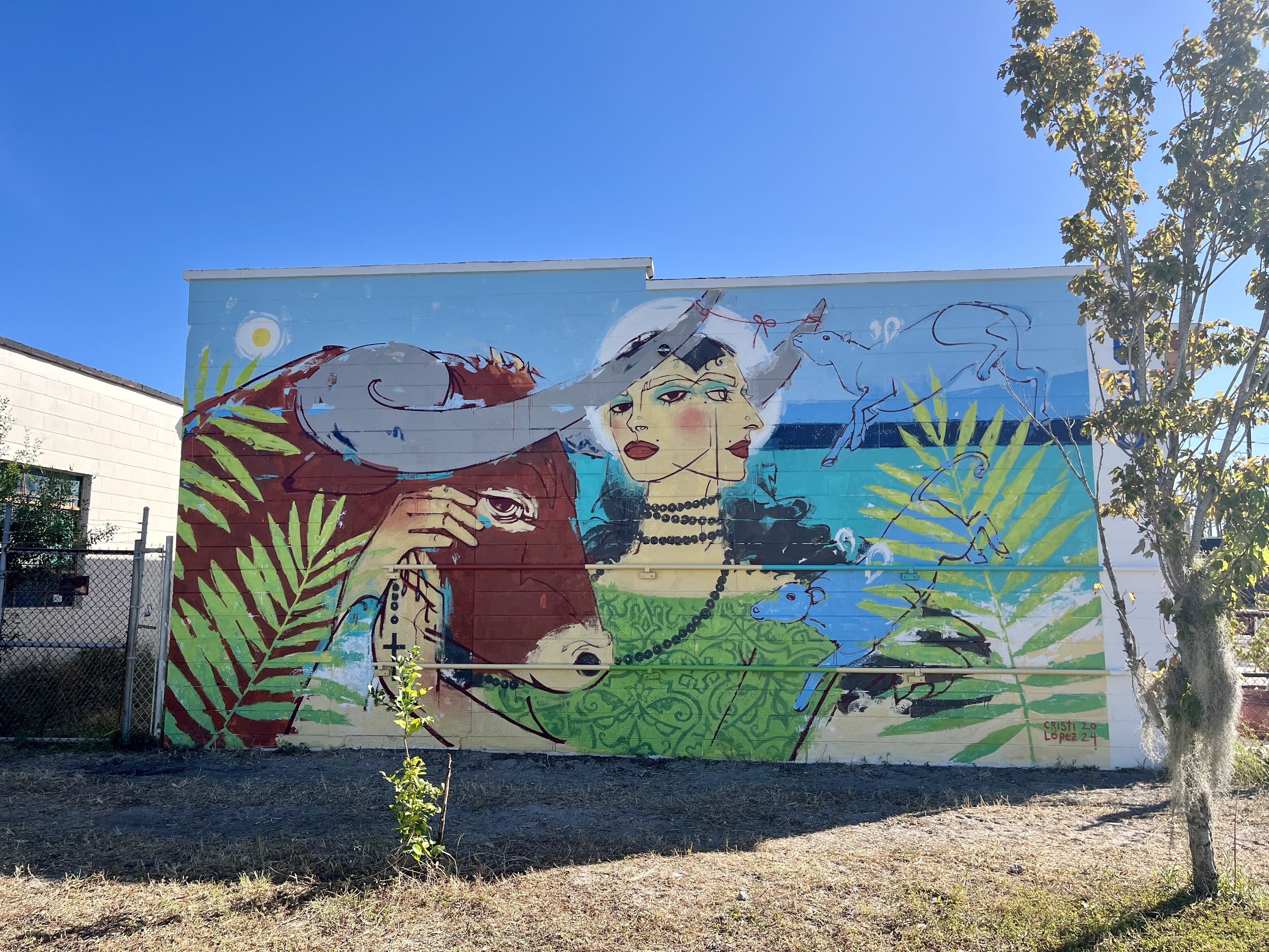 A mural of a woman with black hair and black beads looped around her neck looking at the viewer with her hand caressing a brown bull. The mural is decorated with palm fronds.