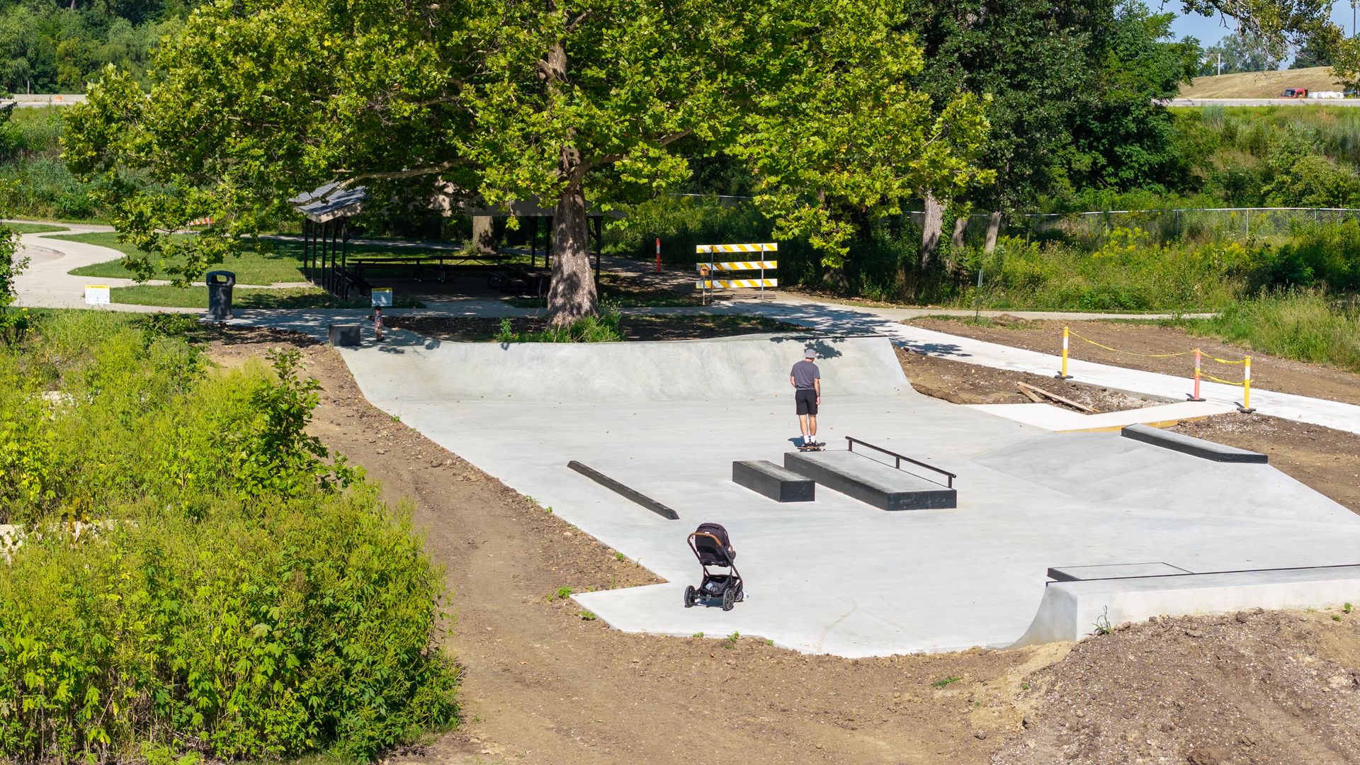 Sunny park with a large central tree over a concrete skate park. A person skateboards on a ramp while a stroller sits nearby; a shelter and fencing are in the background.