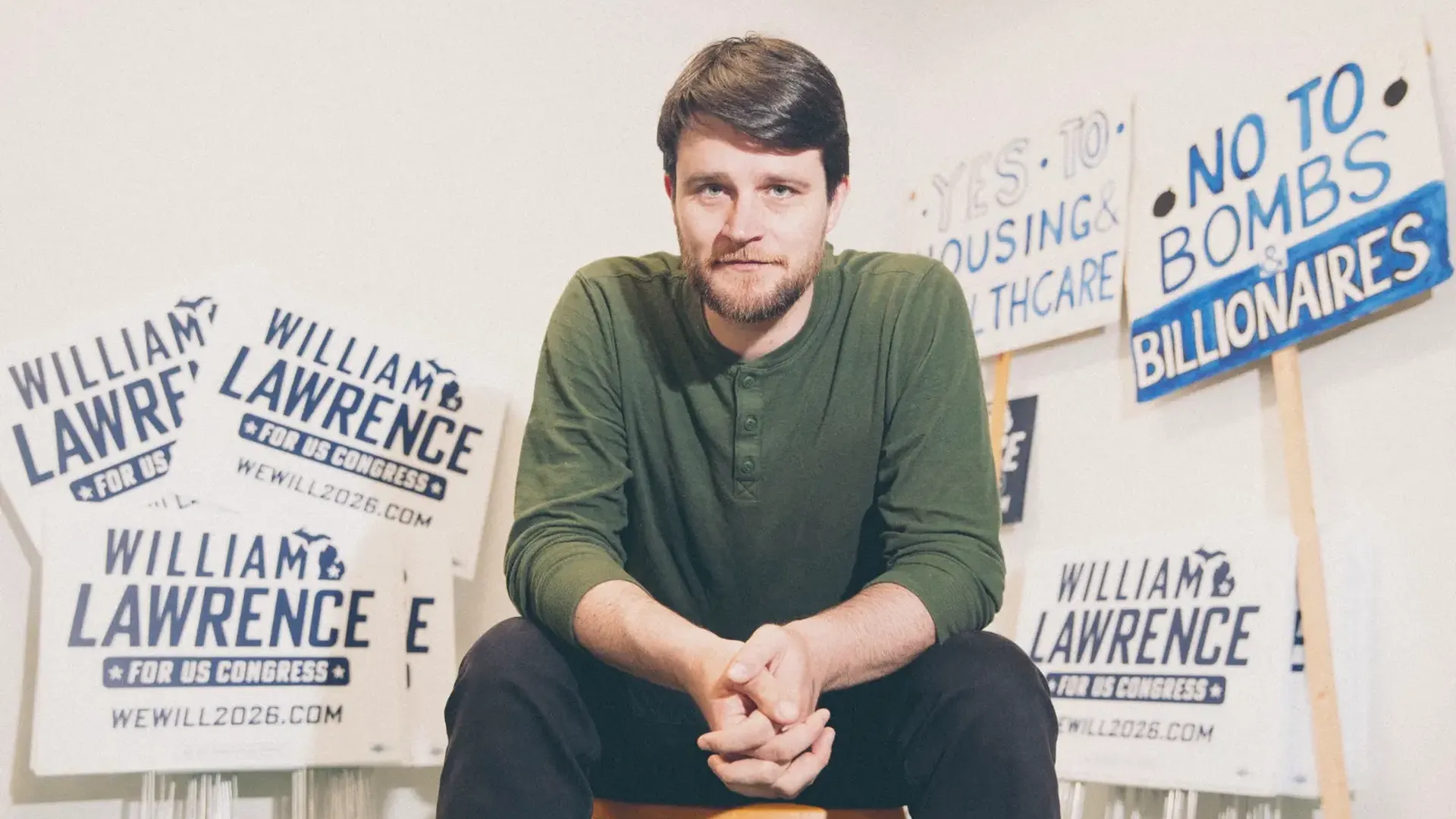 A man with short dark hair and beard sits on a chair in a green shirt, hands clasped. Behind him are campaign signs reading William Lawrence for US Congress and No to bombs, billionaires.