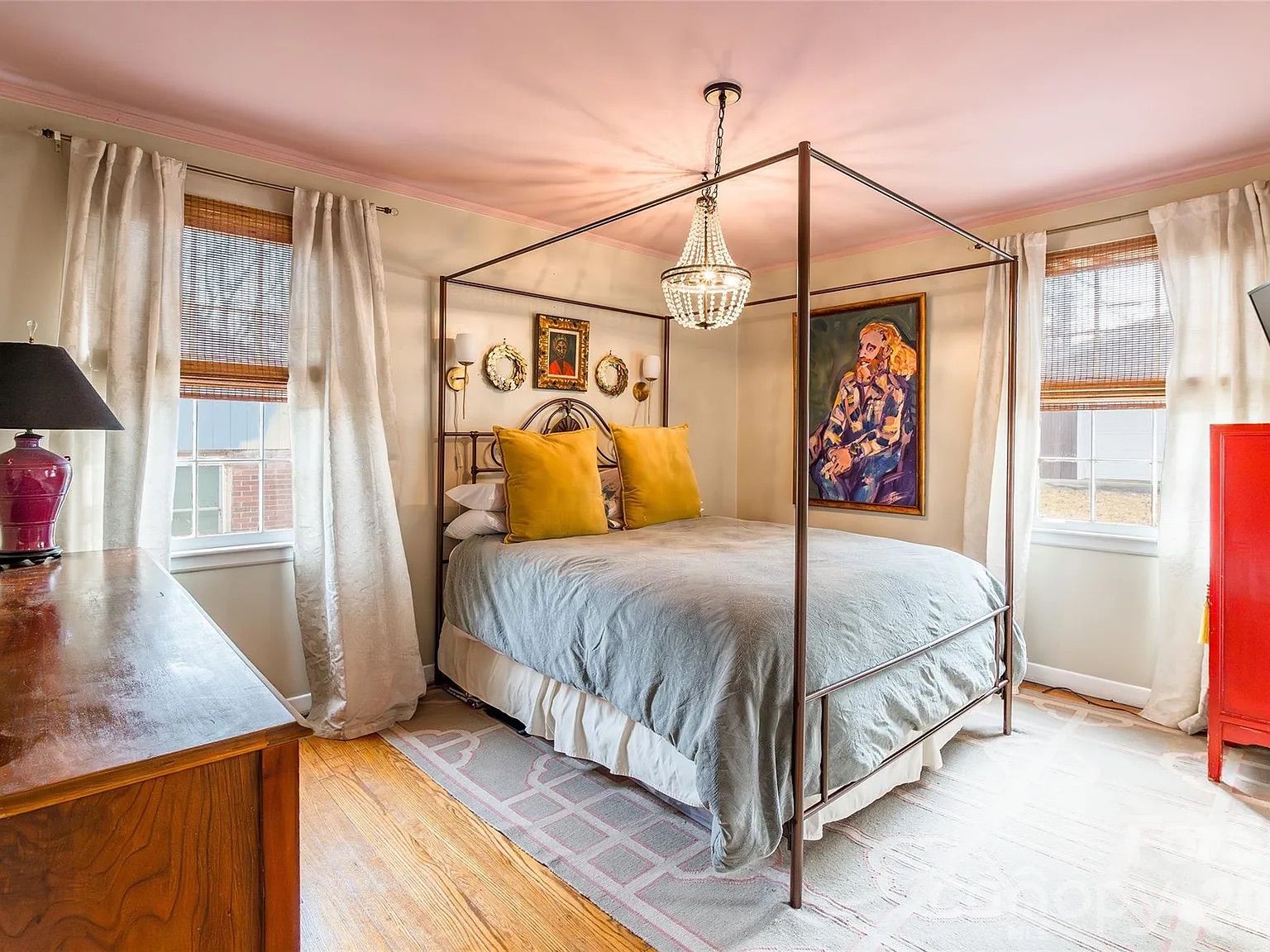 Cozy bedroom with a four-poster metal bed, gray bedding, two large mustard yellow pillows, two windows with white curtains and bamboo blinds, a chandelier, colorful wall art, and wooden furniture.